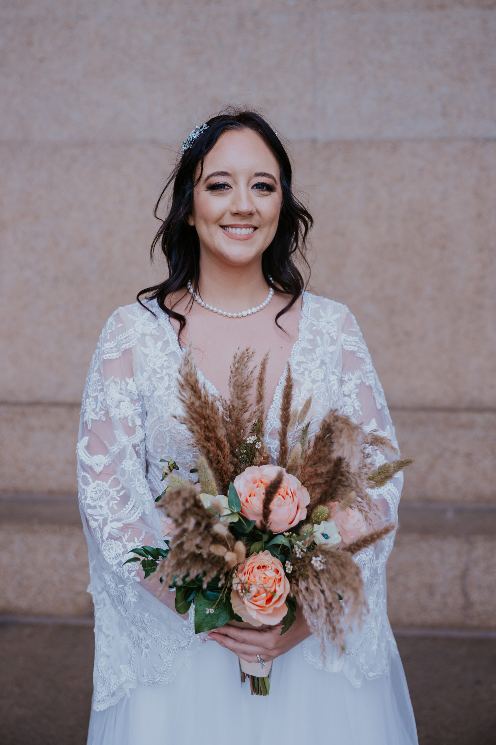 Bride smiling while holding a peach and pampas bouquet, wearing a lace gown and pearl necklace in front of a stone wall.