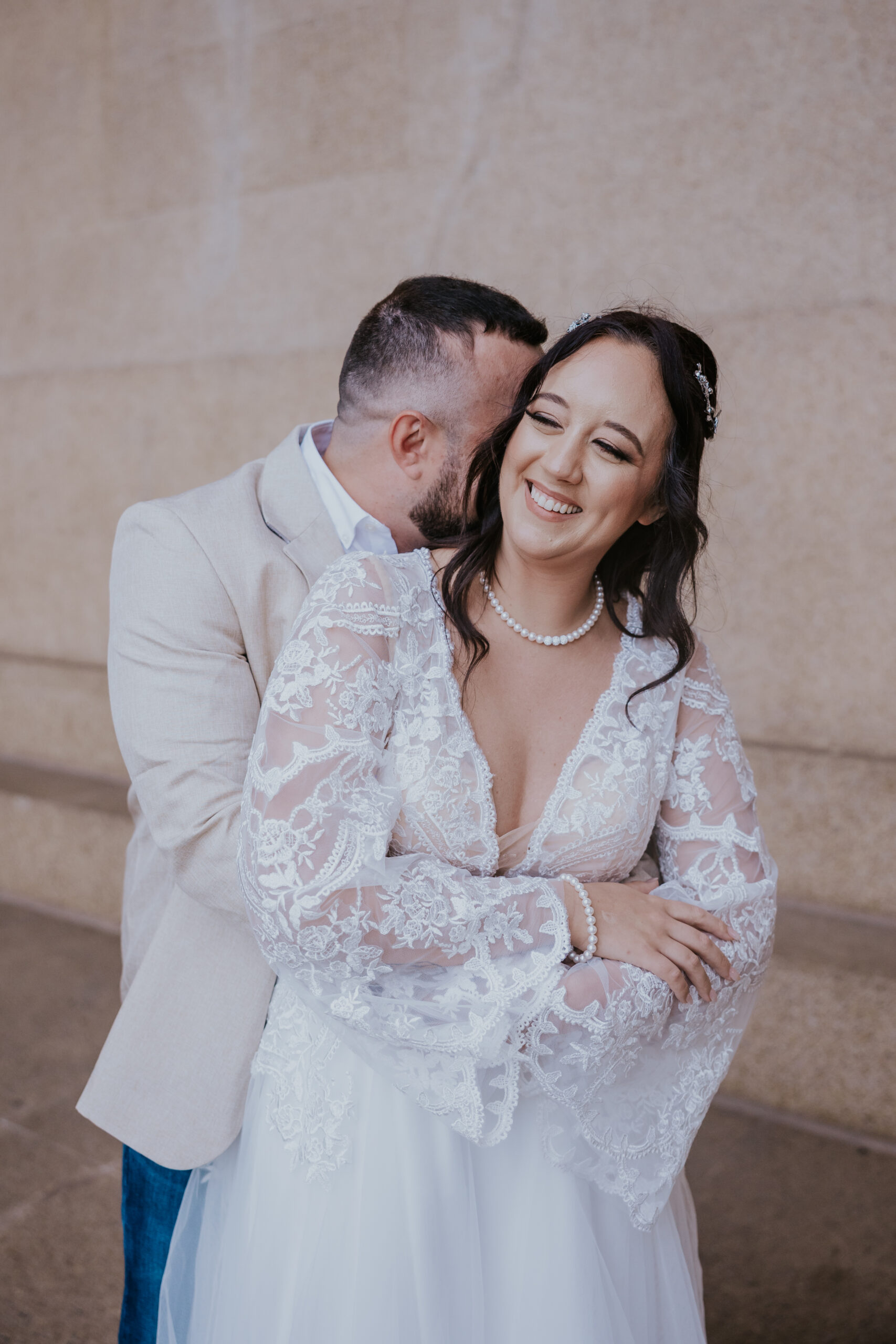 Candid portrait of bride smiling while groom embraces her from behind in front of a stone wall.
