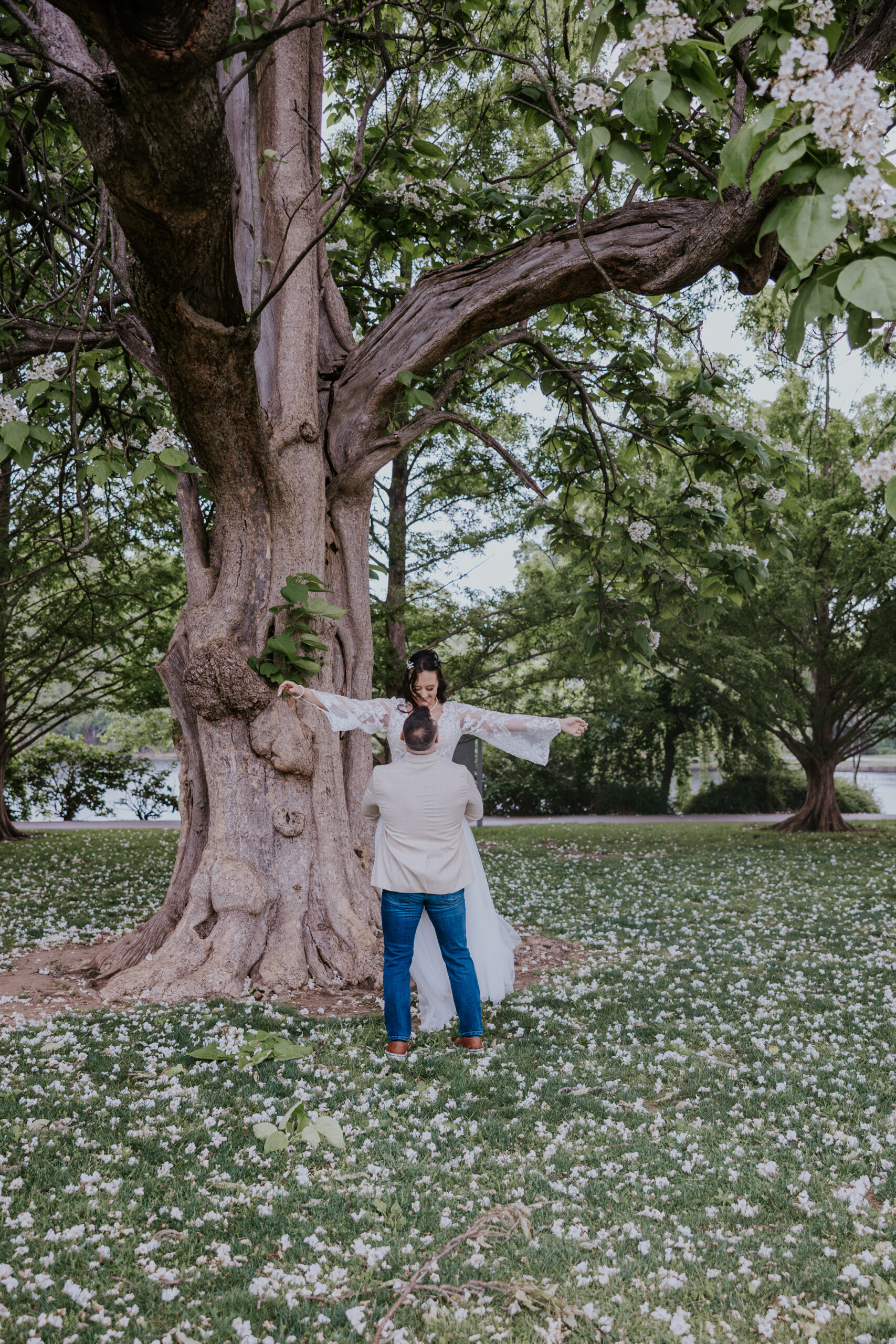 Bride extending her arms as groom lifts her in front of a large tree covered in white blossoms—whimsical wedding portrait.