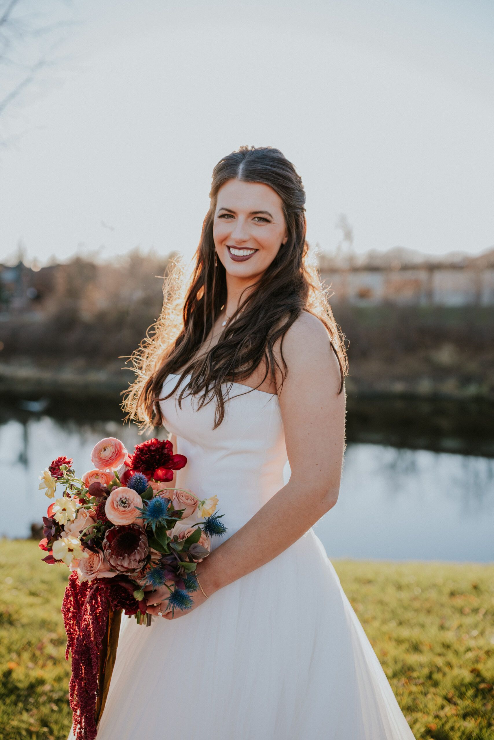 Bride standing by the water in a classic strapless gown, holding a vibrant bouquet and glowing in the sunset.
