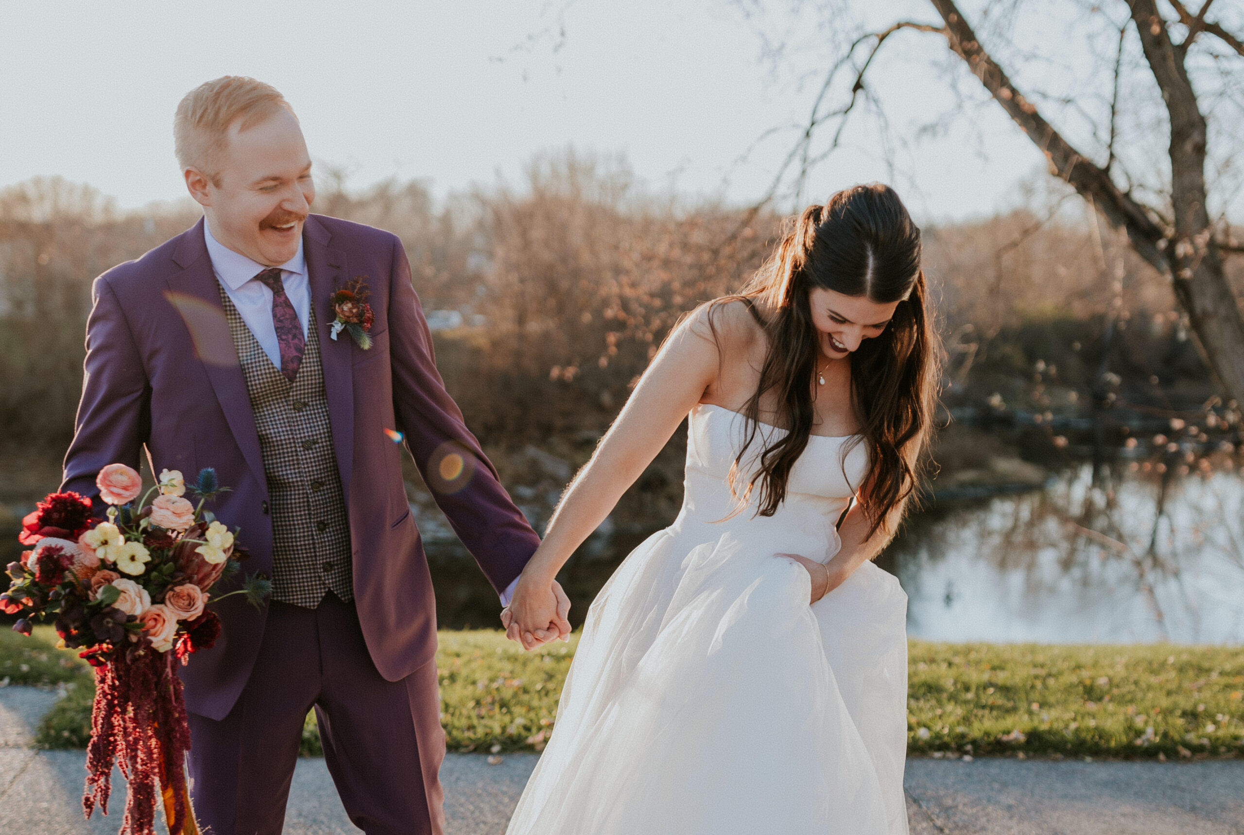 Bride and groom laughing while walking near a quiet lake at golden hour, holding hands with joy.