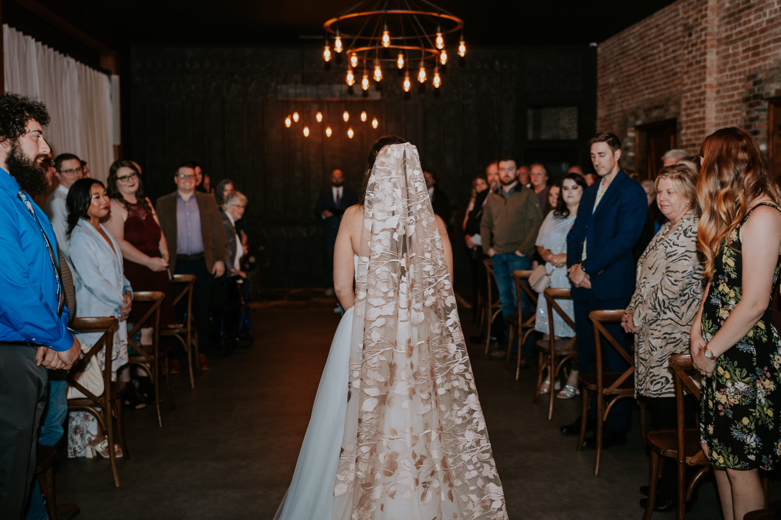 Bride walking down the aisle with a floral veil as guests look on, the ceremony space lit with modern chandeliers.