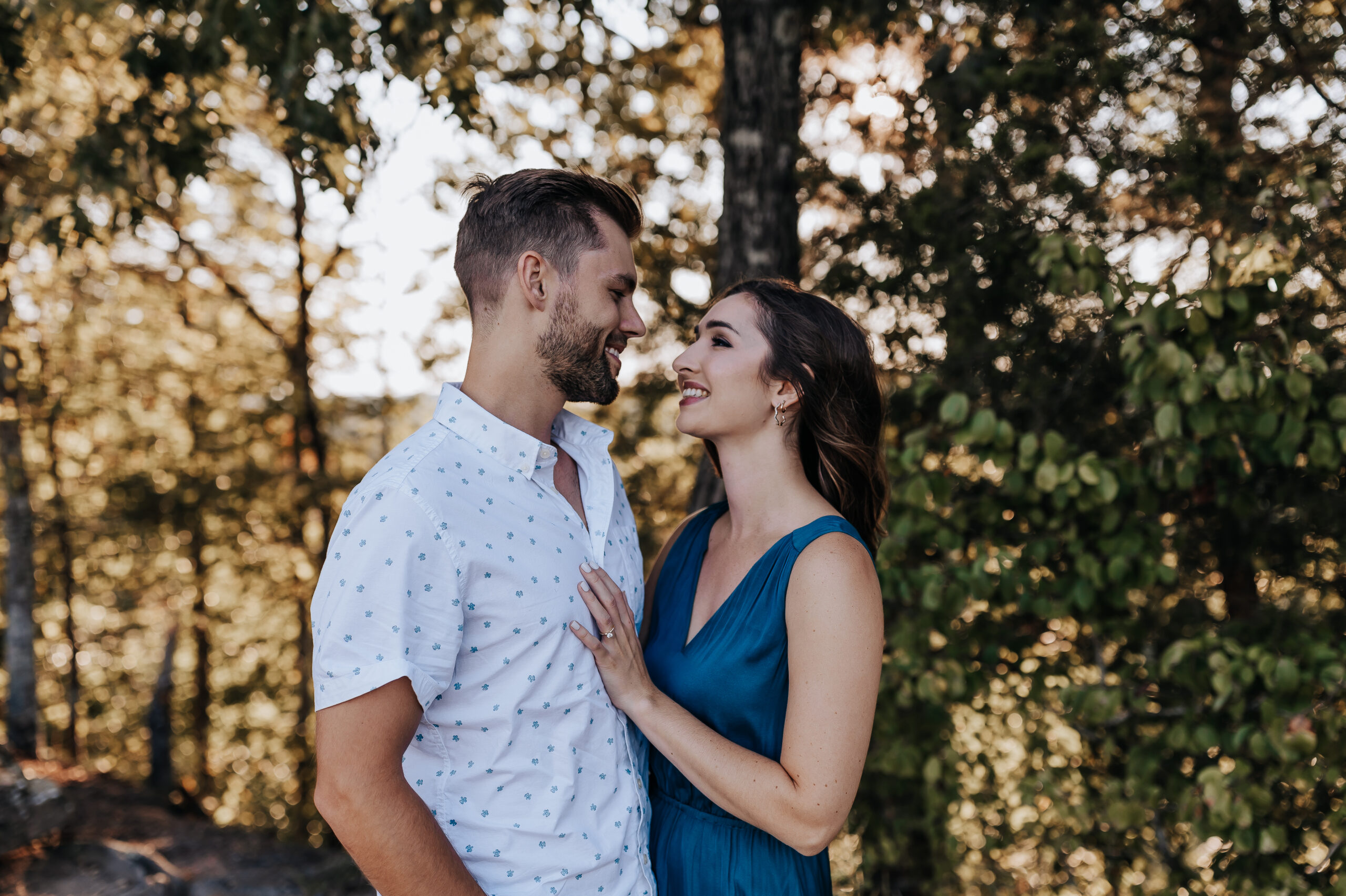 A couple stands close in a wooded area, smiling and sharing a soft gaze while sunlight filters through the trees.