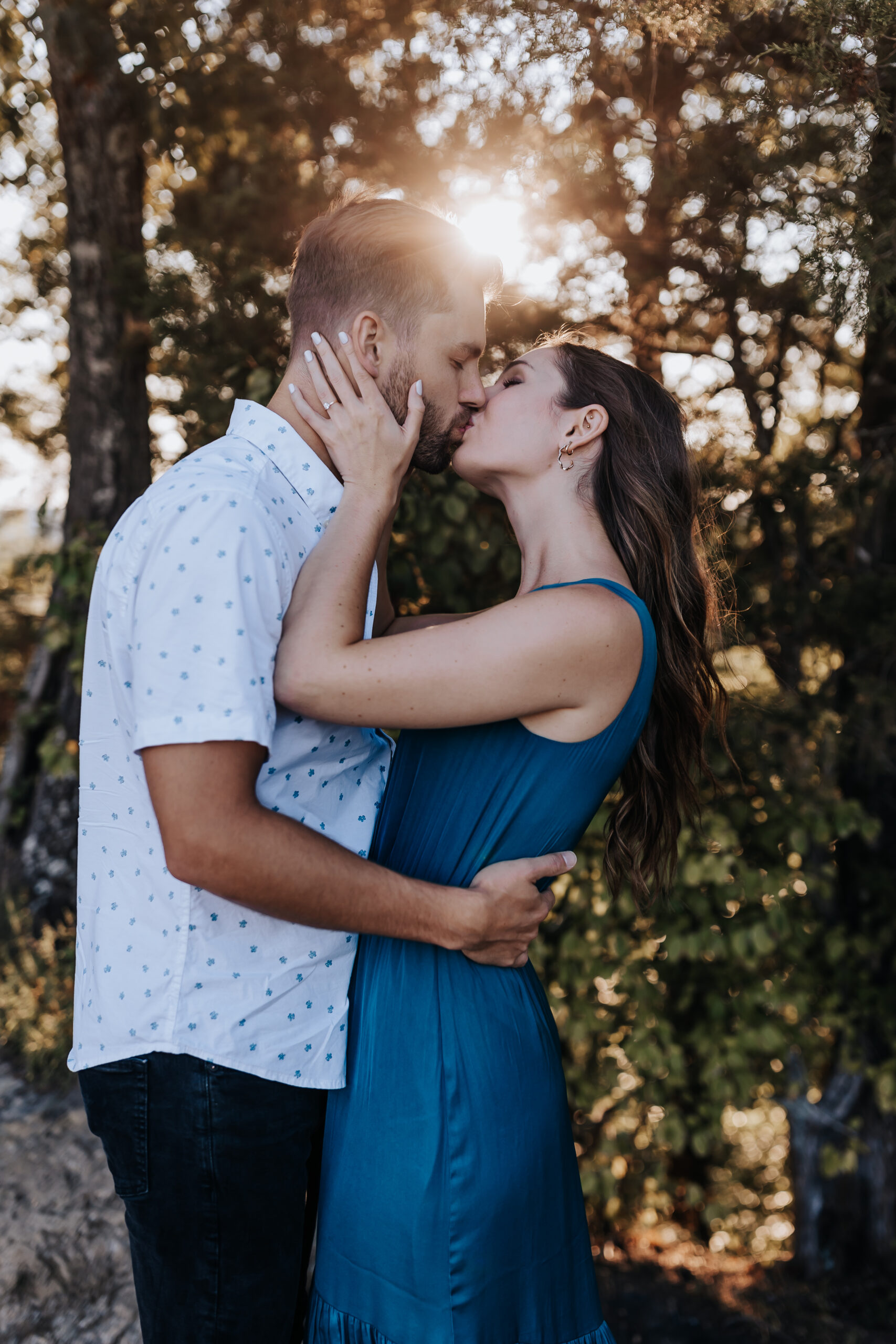 A couple shares a romantic kiss at sunset, light shining through the trees behind them, captured by a Nashville engagement photographer.