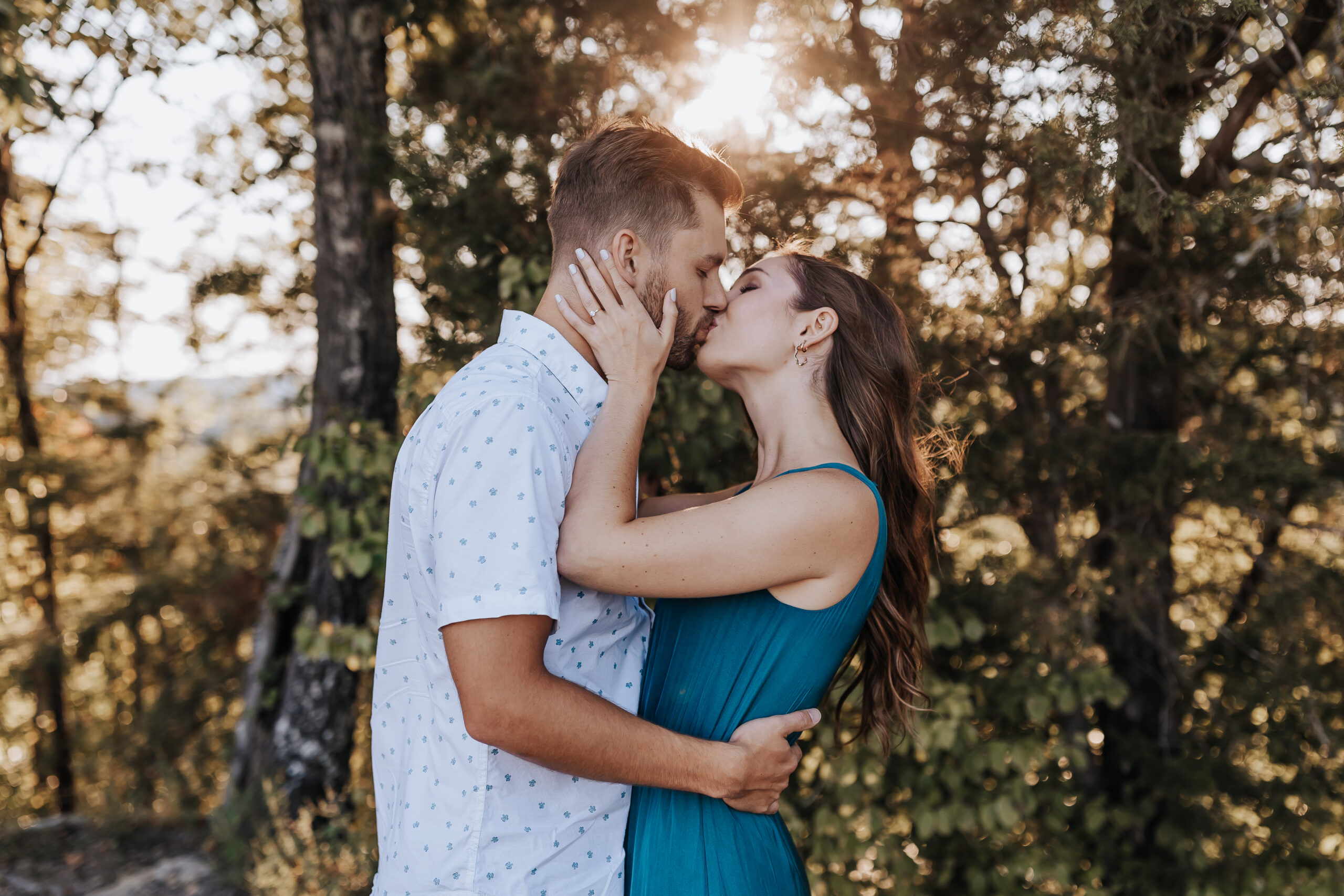 A romantic kiss shared beneath the trees as sunlight glows behind the couple, creating a dreamy and intimate forest moment.