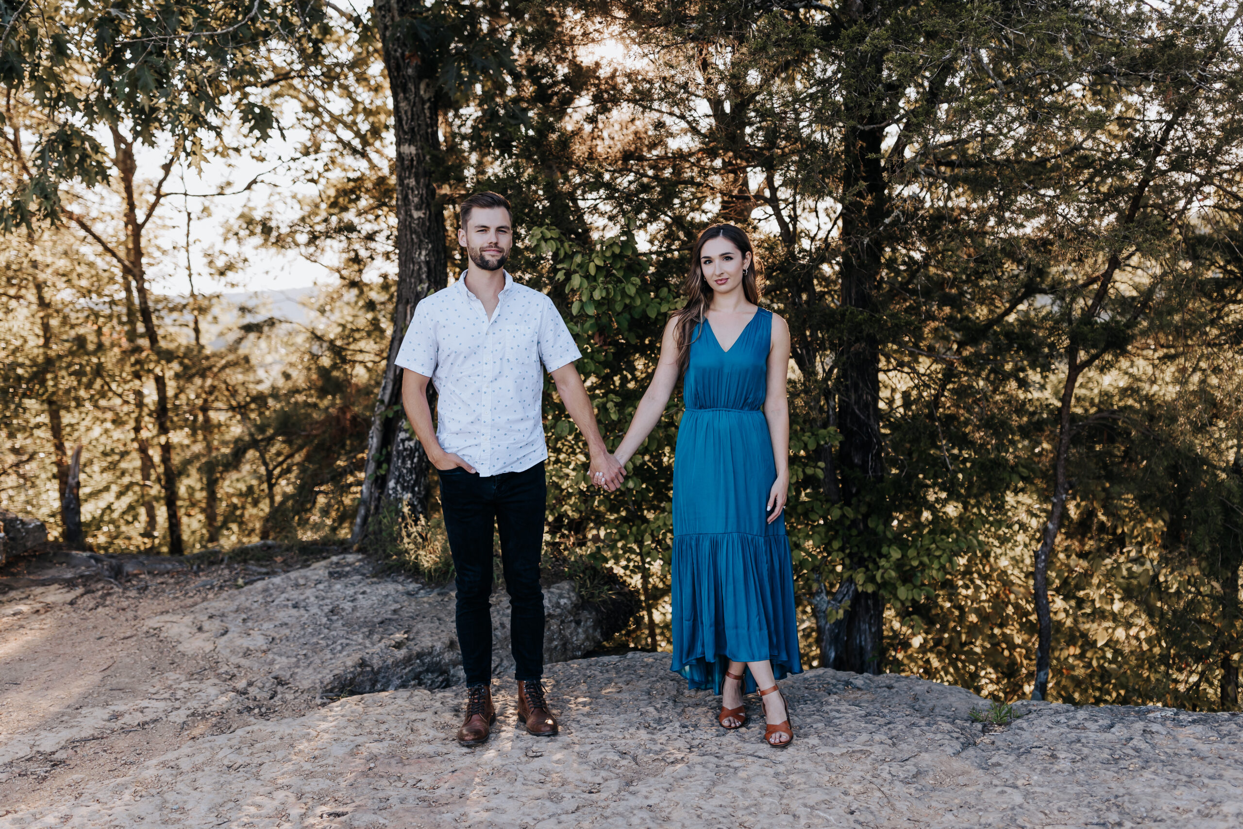 A couple holds hands and faces the camera while standing on a rocky overlook surrounded by tall trees.