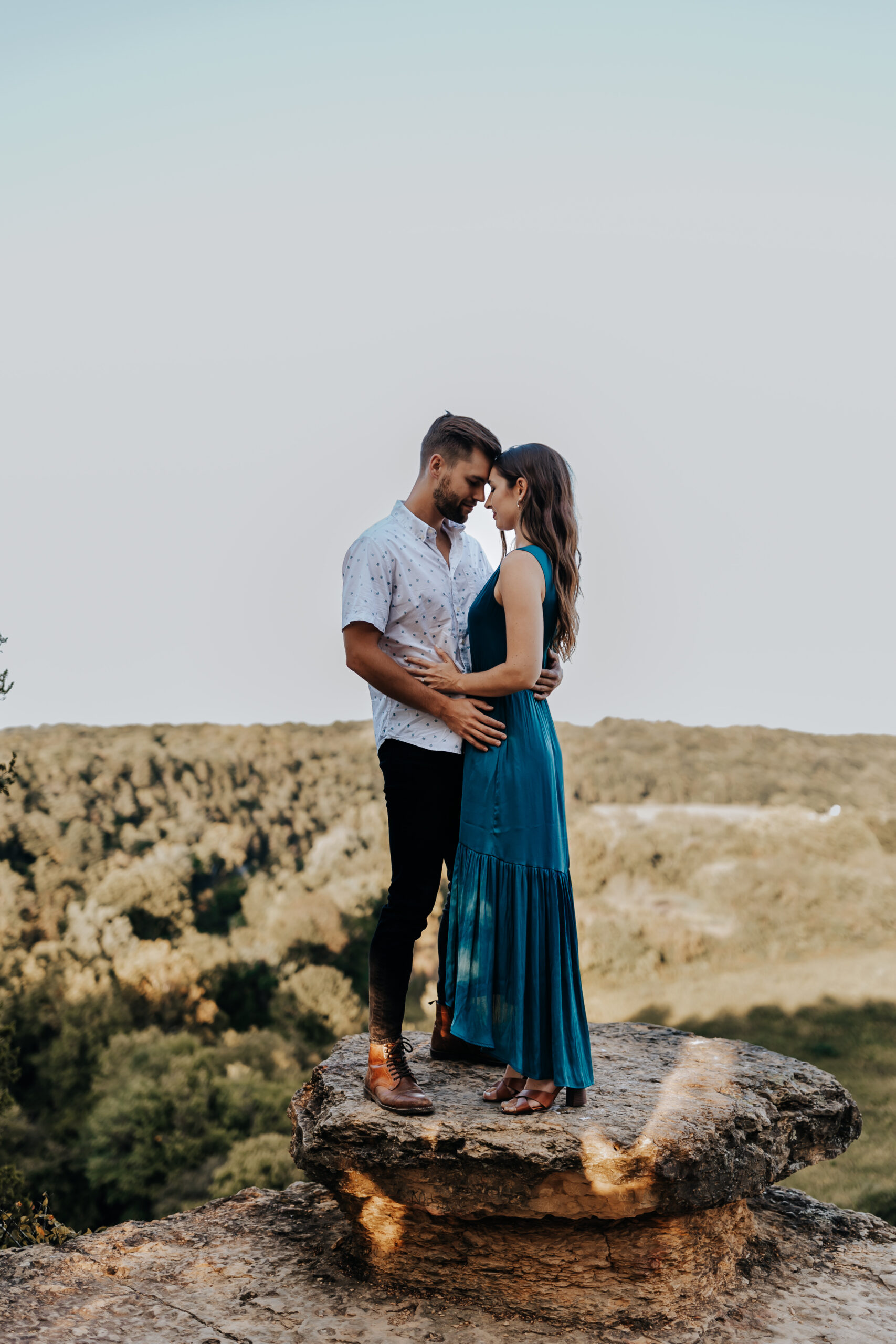 A couple stands on a scenic cliff edge, foreheads touching, with a vast view of rolling hills behind them during their session with a Nashville engagement photographer.