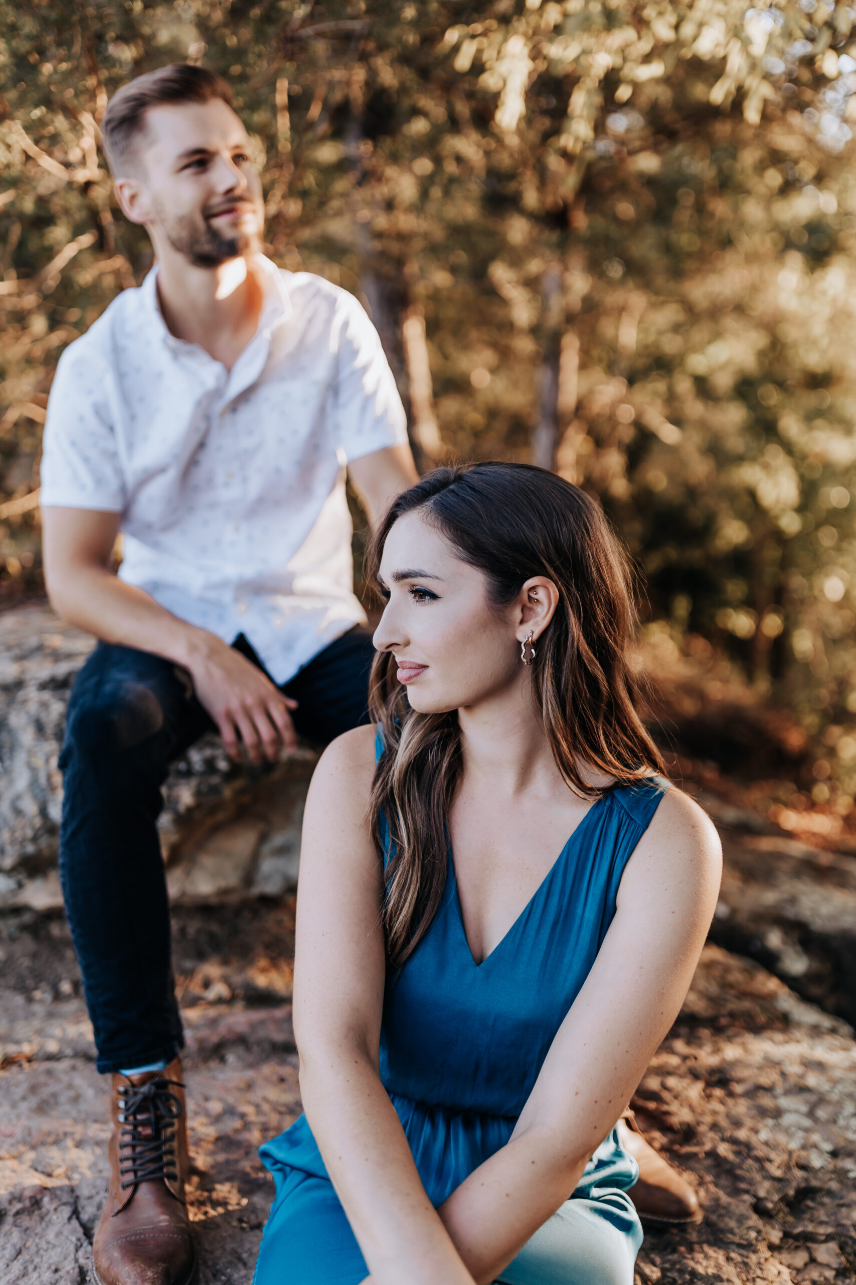 A couple sits together on a rocky path surrounded by trees during golden hour, sharing a quiet, reflective moment.