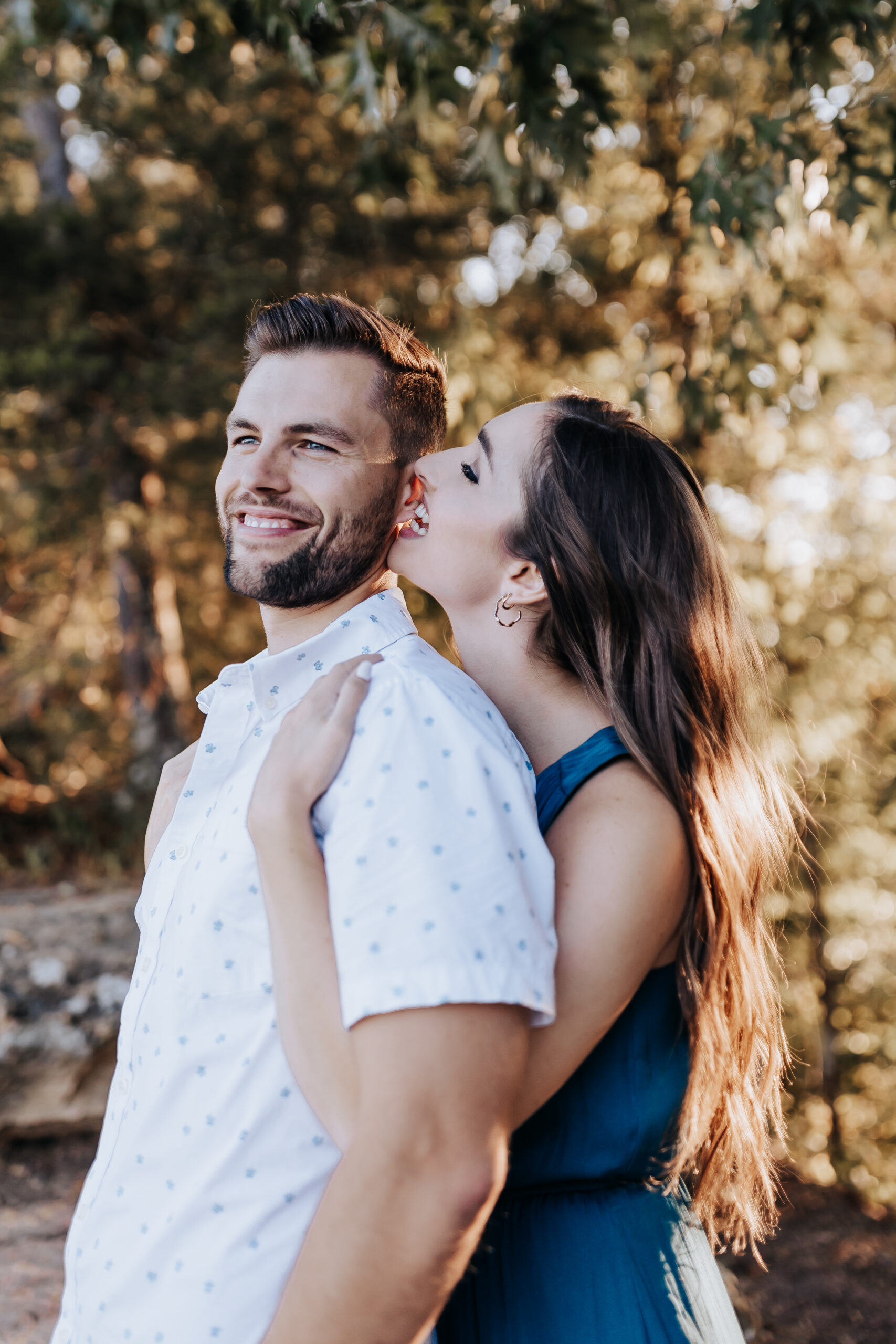 A playful moment as a woman kisses her partner on the cheek while standing close in a wooded setting, captured by a Nashville engagement photographer.