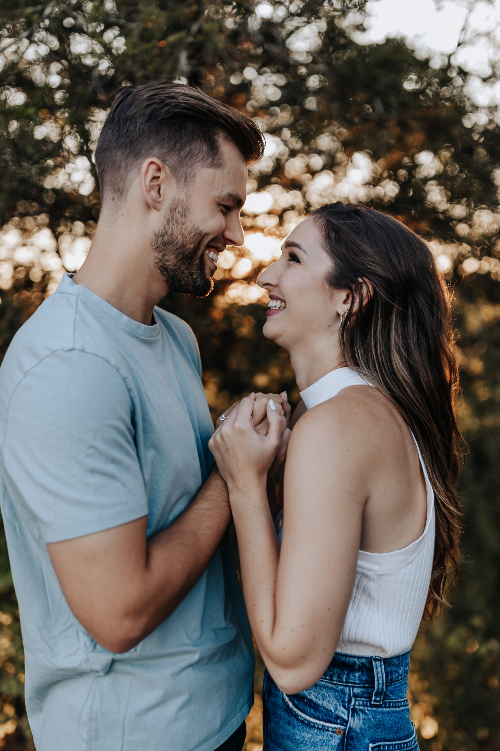 A joyful couple stands face to face, smiling and holding hands as they enjoy a quiet forest moment together.