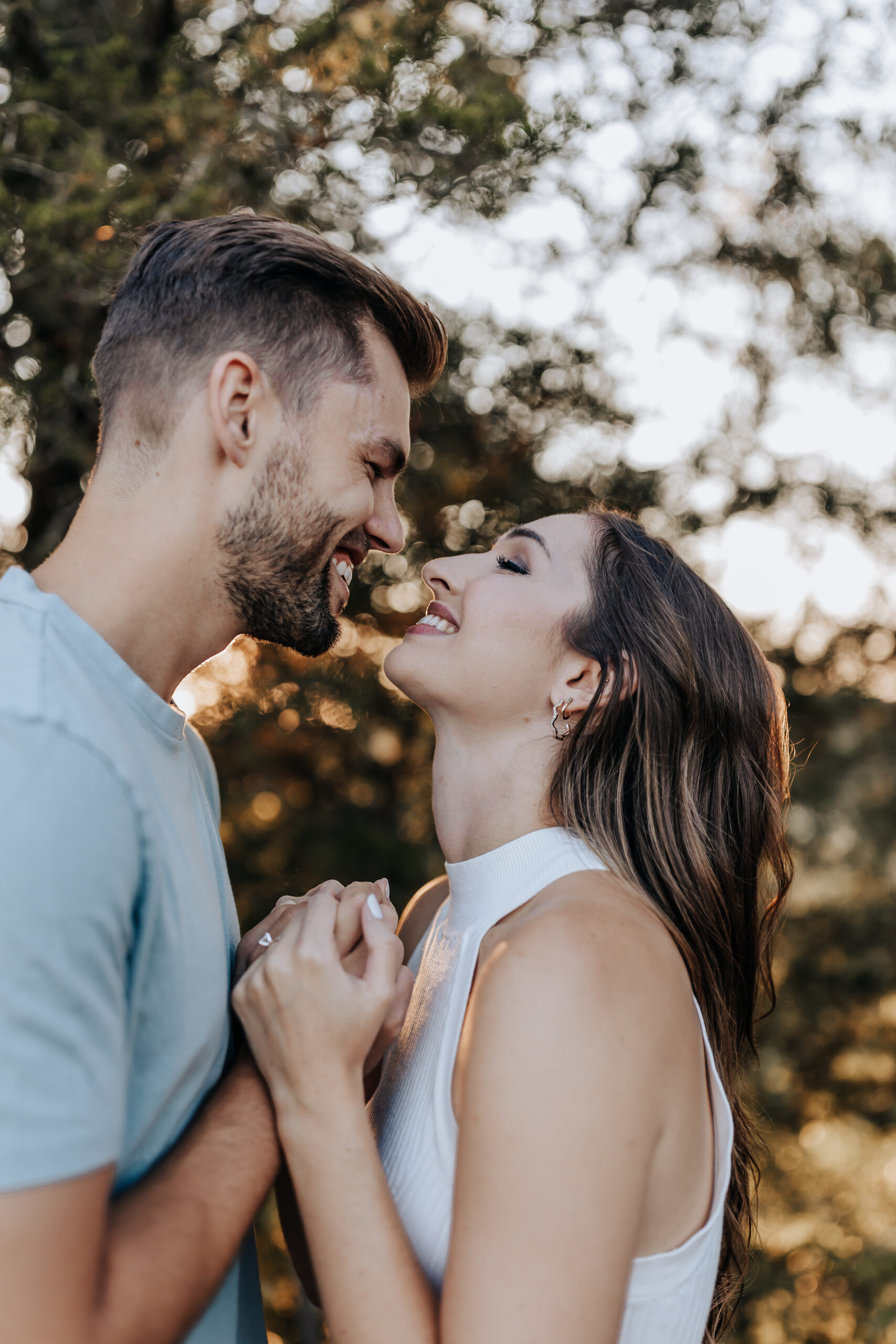 Close-up of a joyful couple leaning in for a kiss while holding hands, sunlight filtering through the trees in the background.