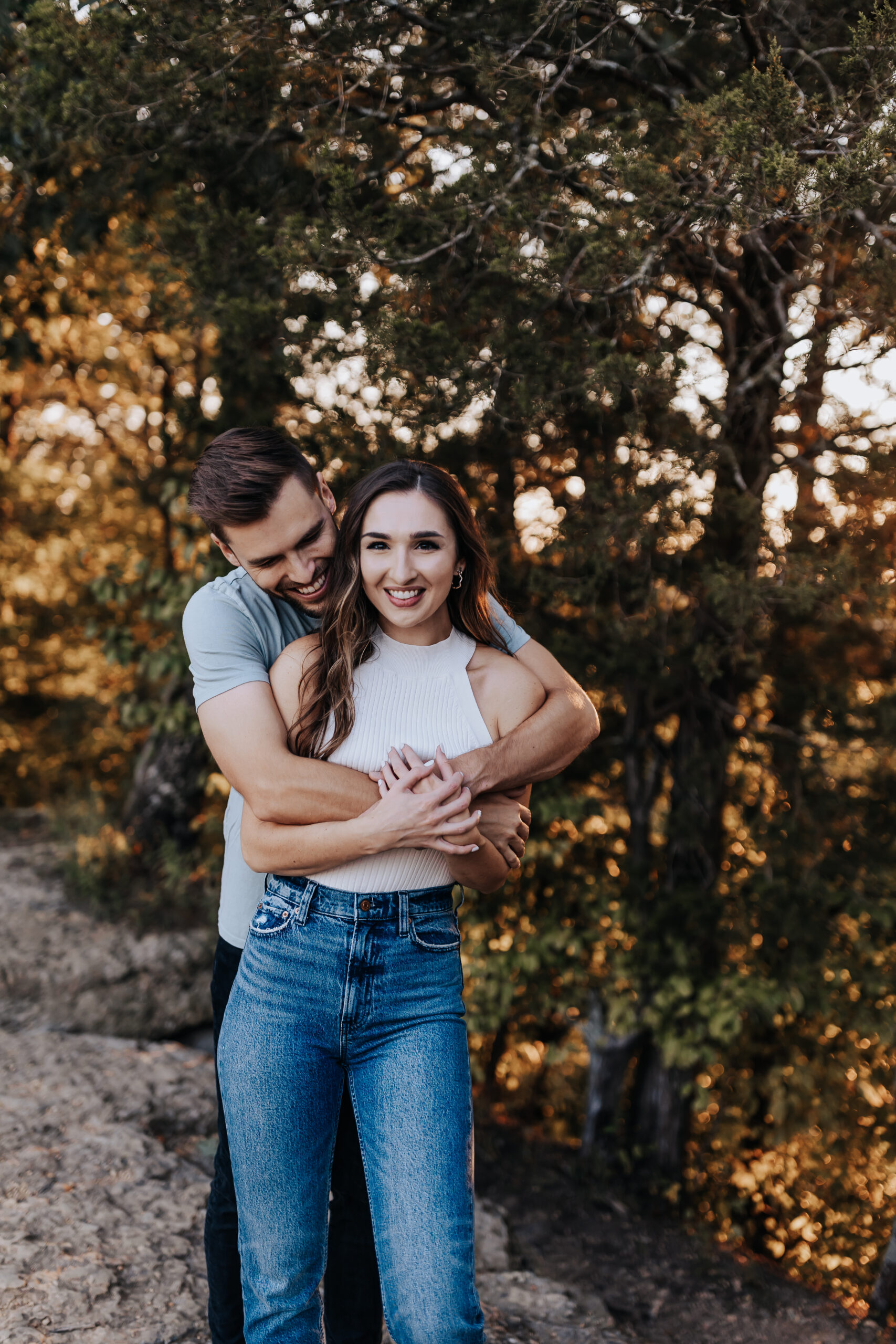 A woman smiles softly while her partner wraps his arms around her from behind during a warm, golden-hour engagement session.