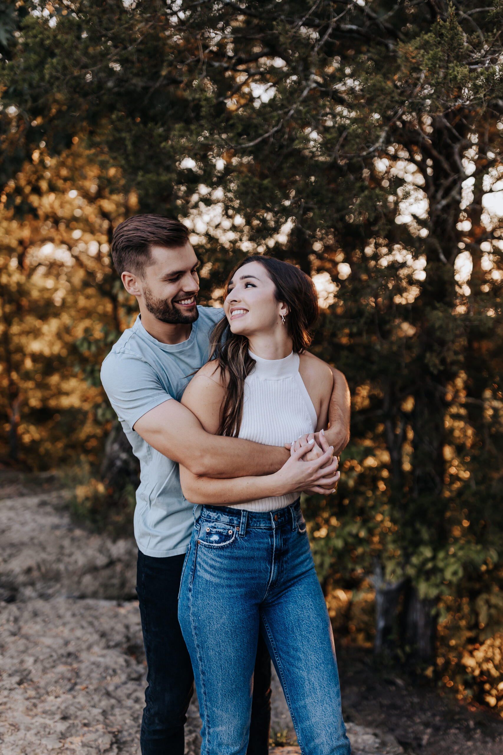 A man wraps his arms around his fiancée as they smile at each other beneath the trees, captured by a Nashville engagement photographer.
