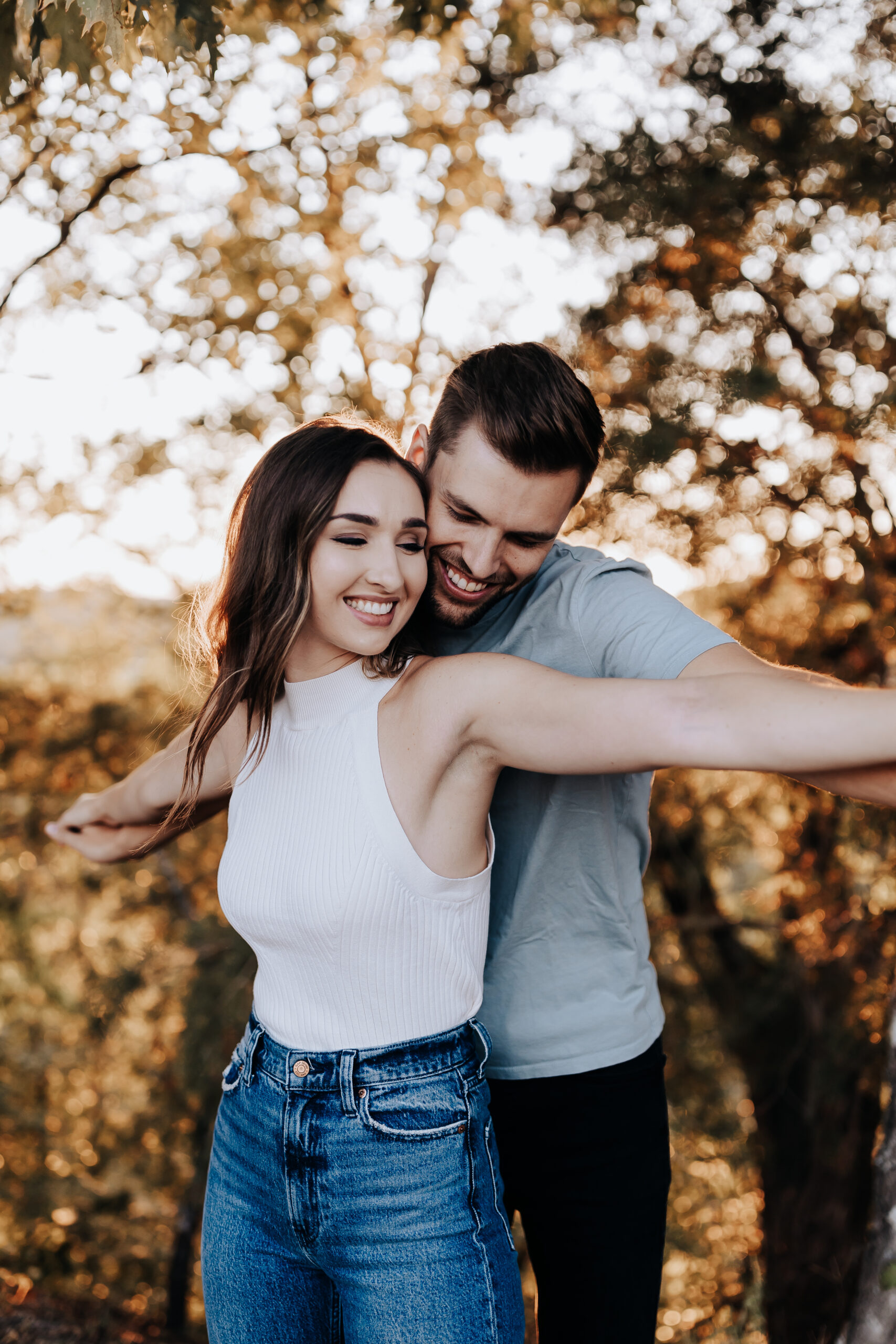 A woman smiles with her arms outstretched as her partner hugs her from behind in a fun, candid moment during their engagement shoot with a Nashville engagement photographer.