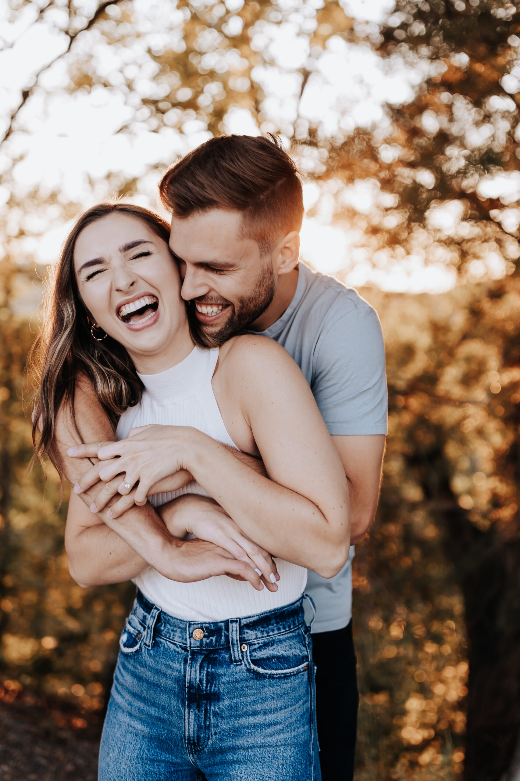 A candid moment of laughter as a woman leans into her partner’s embrace, both beaming with joy in the forest glow.