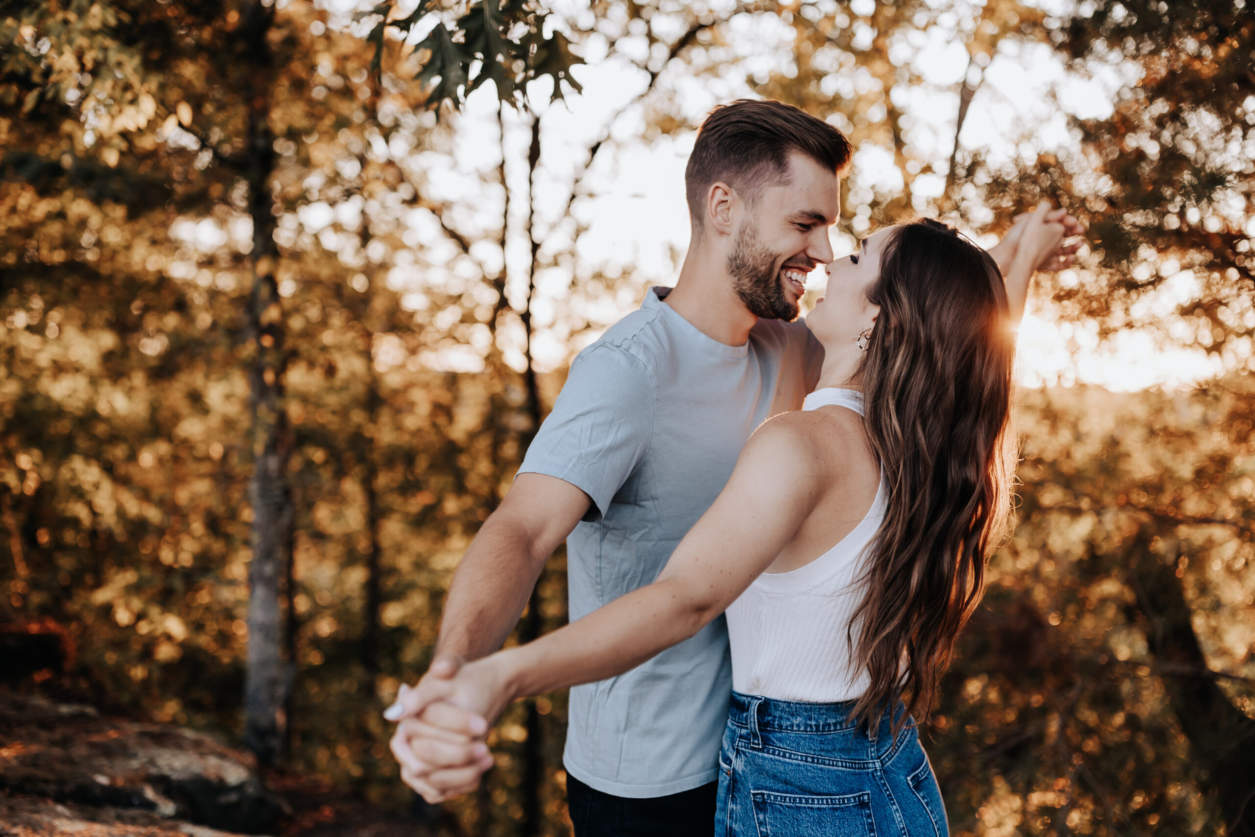 An engaged couple dances joyfully in the forest at sunset, holding hands and laughing together in a romantic golden-hour glow.