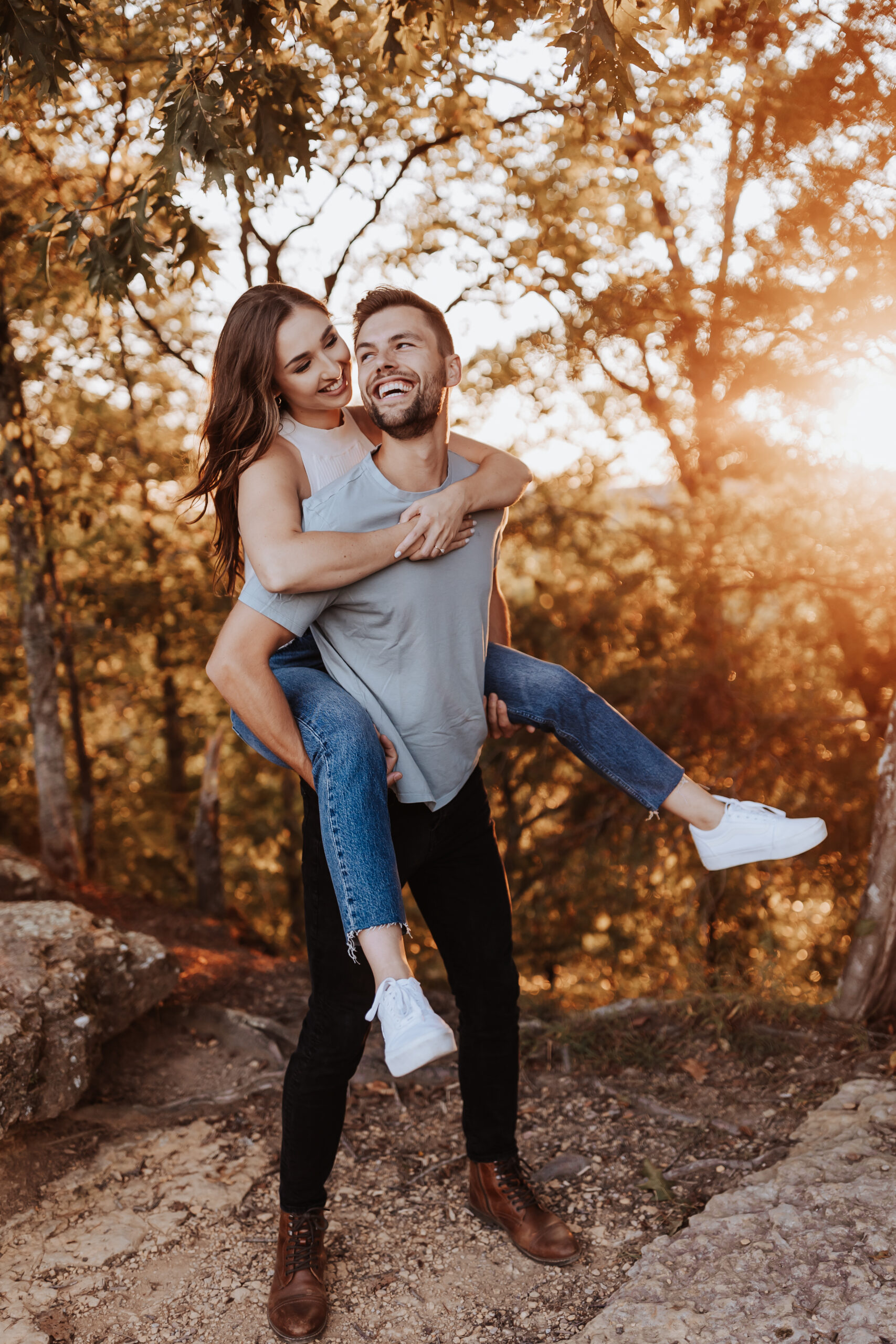 A joyful piggyback ride as golden sunlight filters through the forest, highlighting the couple’s fun and carefree vibe.