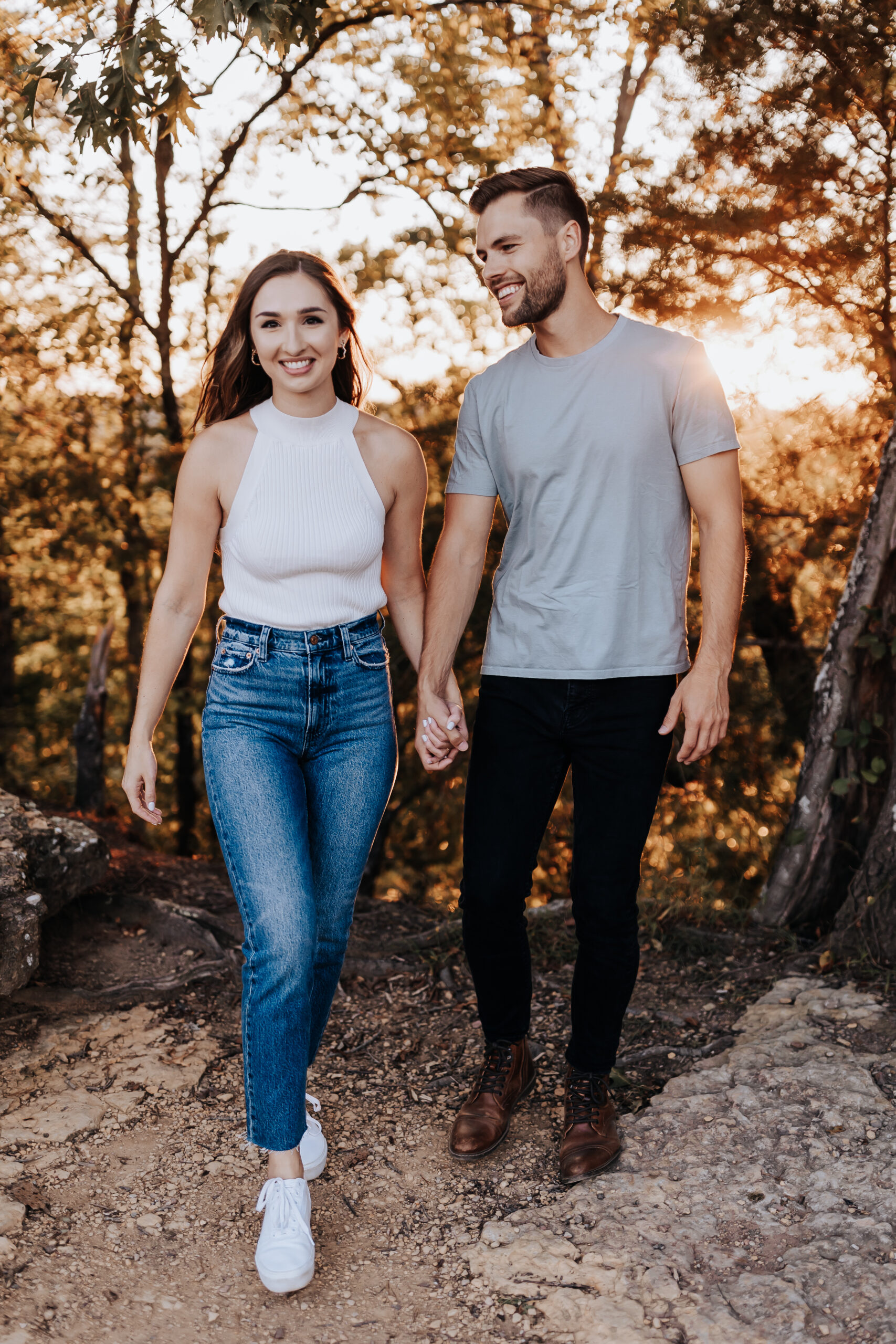 A couple walking hand-in-hand through a wooded trail during golden hour, smiling and relaxed during their session with a Nashville engagement photographer.
