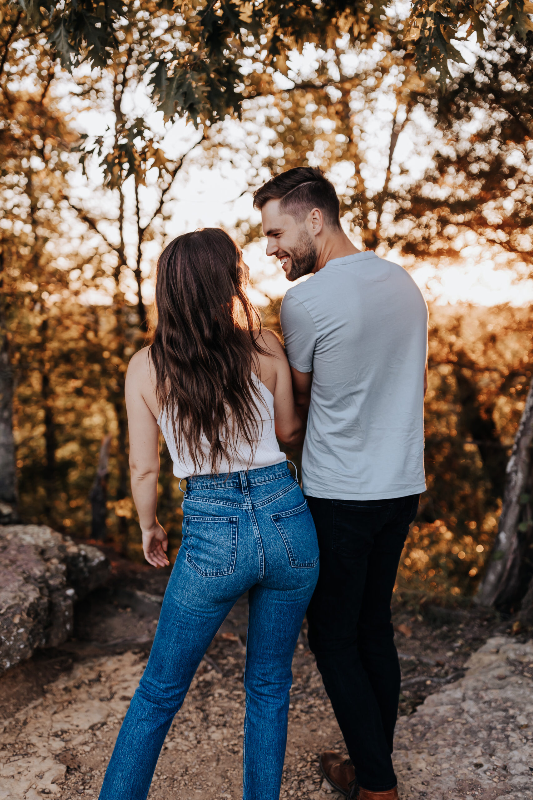 A woman walks slightly ahead of her partner on a forest trail, both smiling at each other under a canopy of golden trees.