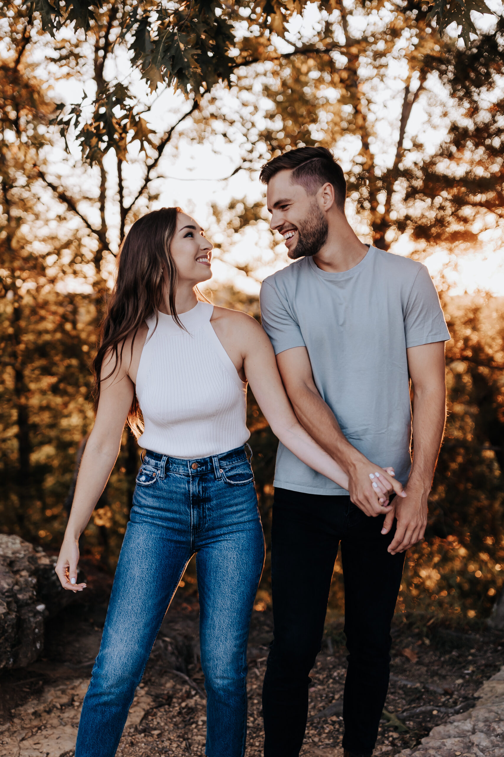 A couple holds hands and walks through a wooded path, smiling at each other in the warm light of sunset.