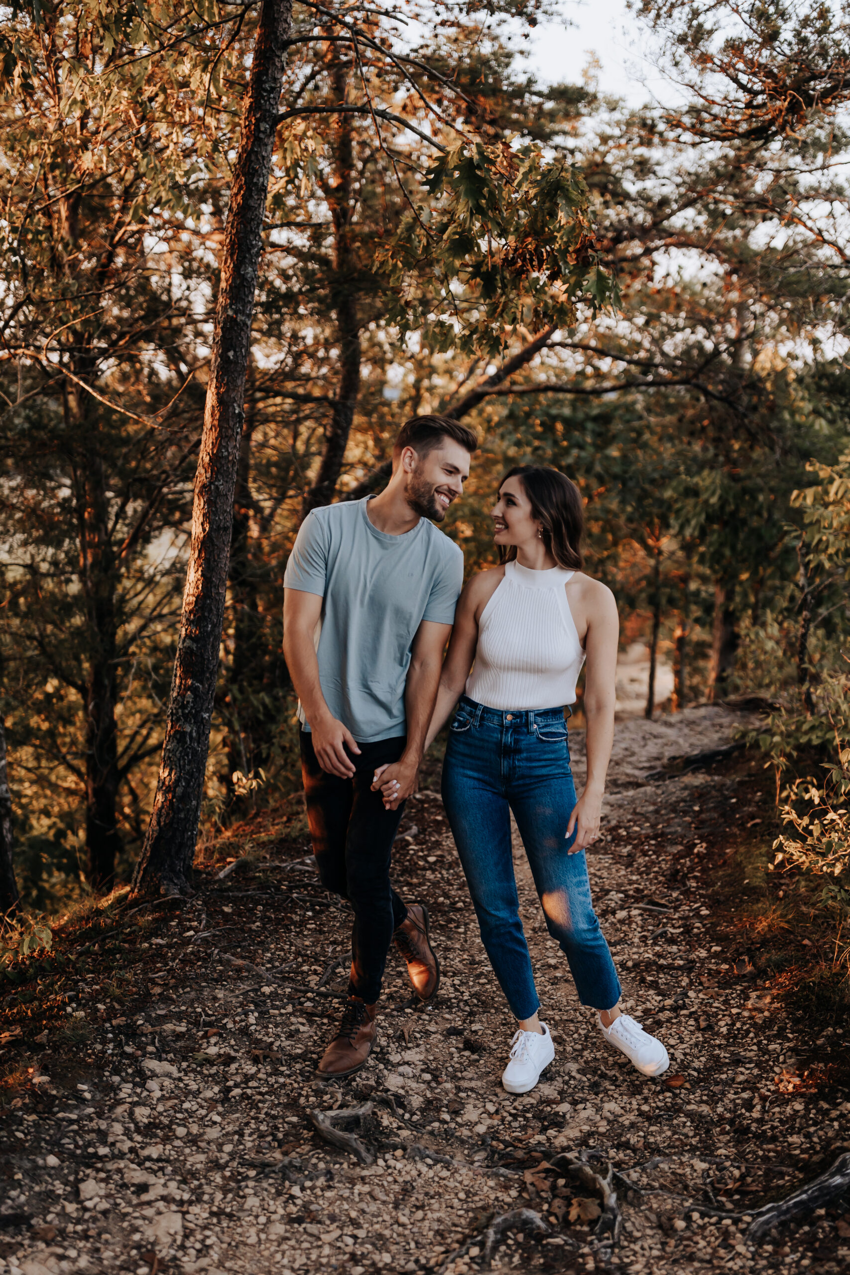 A joyful couple walks hand-in-hand down a forest path at sunset, smiling into each other’s eyes.