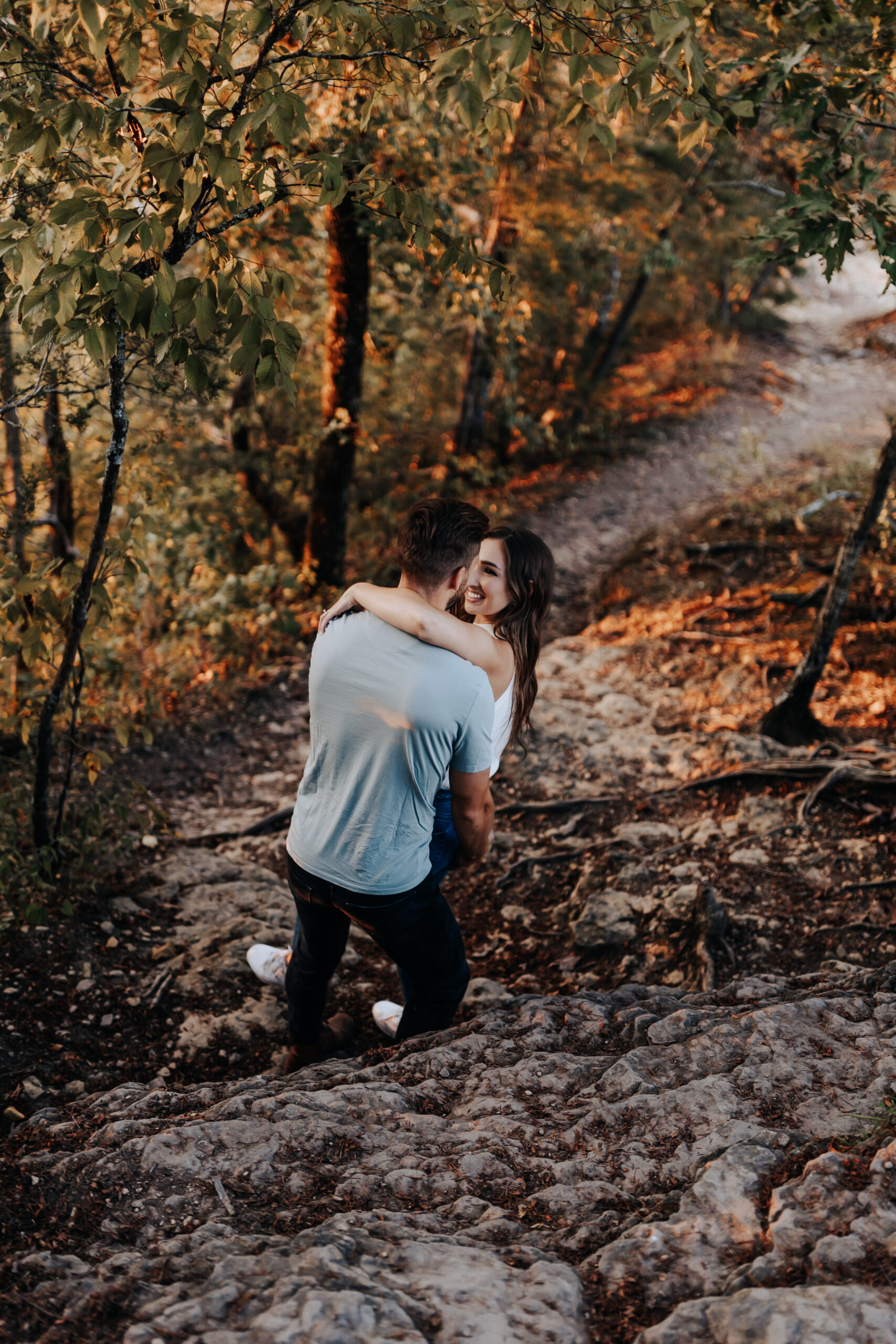 A playful moment as a woman wraps her arms around her fiancé’s shoulders while he carries her up a rocky trail, smiling back at the camera.