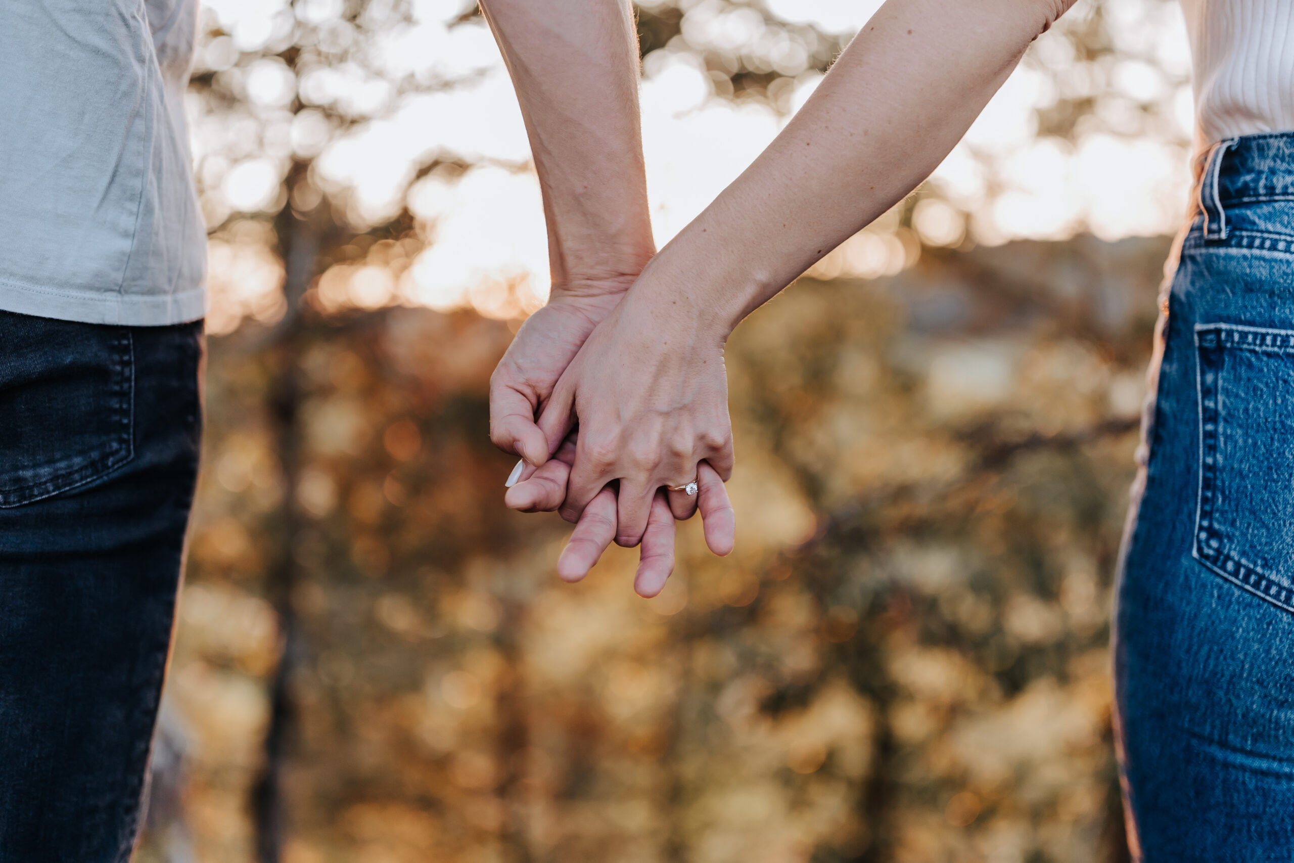 Close-up of a couple holding hands at sunset, with an engagement ring visible as golden light shines through the trees.