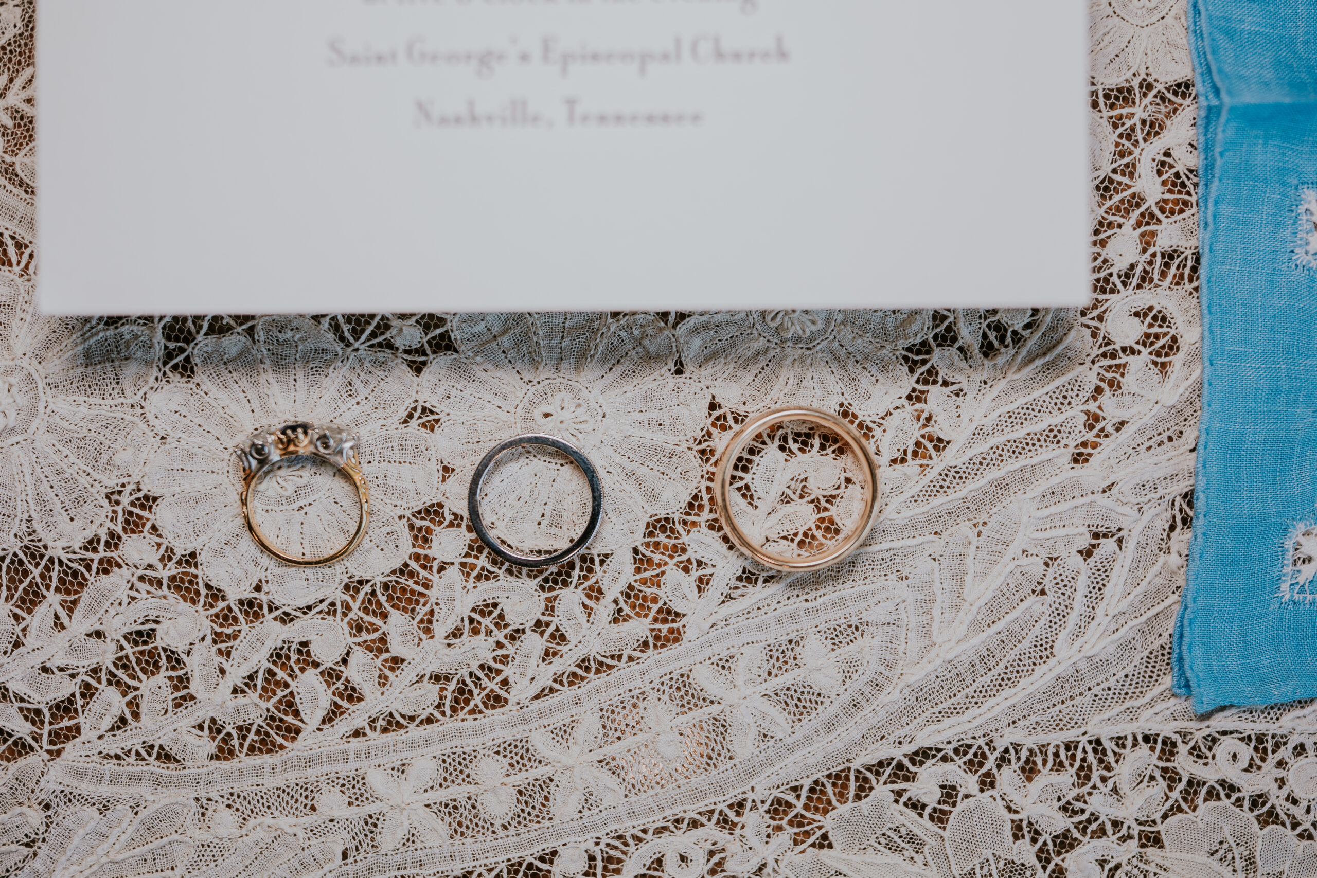 Three wedding bands rest atop delicate lace next to the invitation, showcasing timeless love and elegant details.