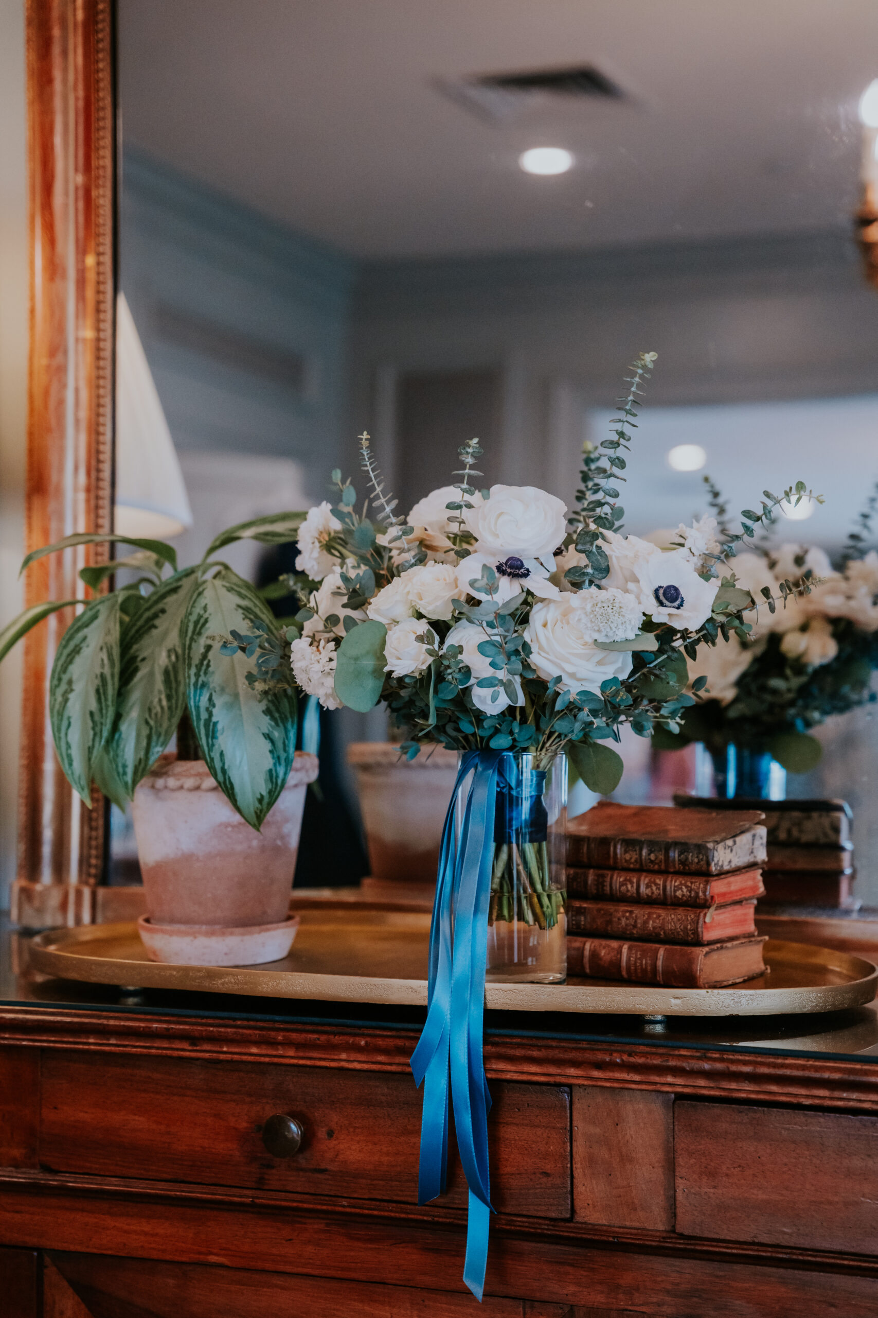 Elegant bridal bouquet of white ranunculus and eucalyptus with a navy ribbon, styled on a vintage dresser with stacked books.