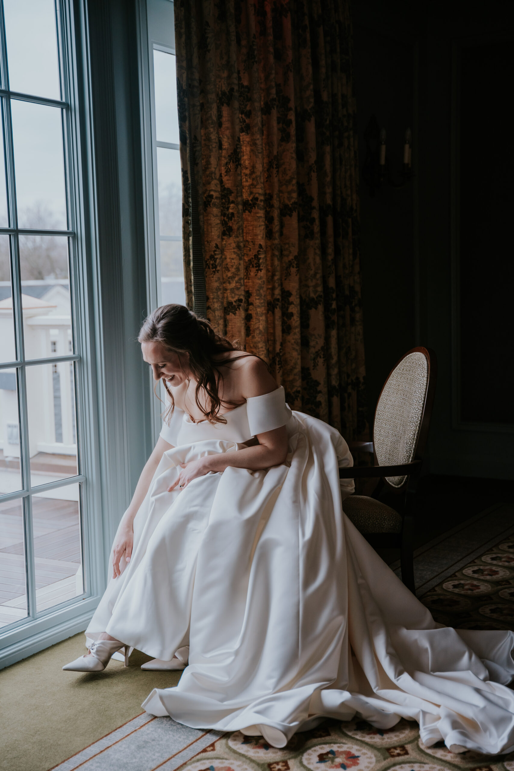 Bride gracefully bends to adjust her heels while sunlight pours in through tall windows, creating a quiet and reflective moment.