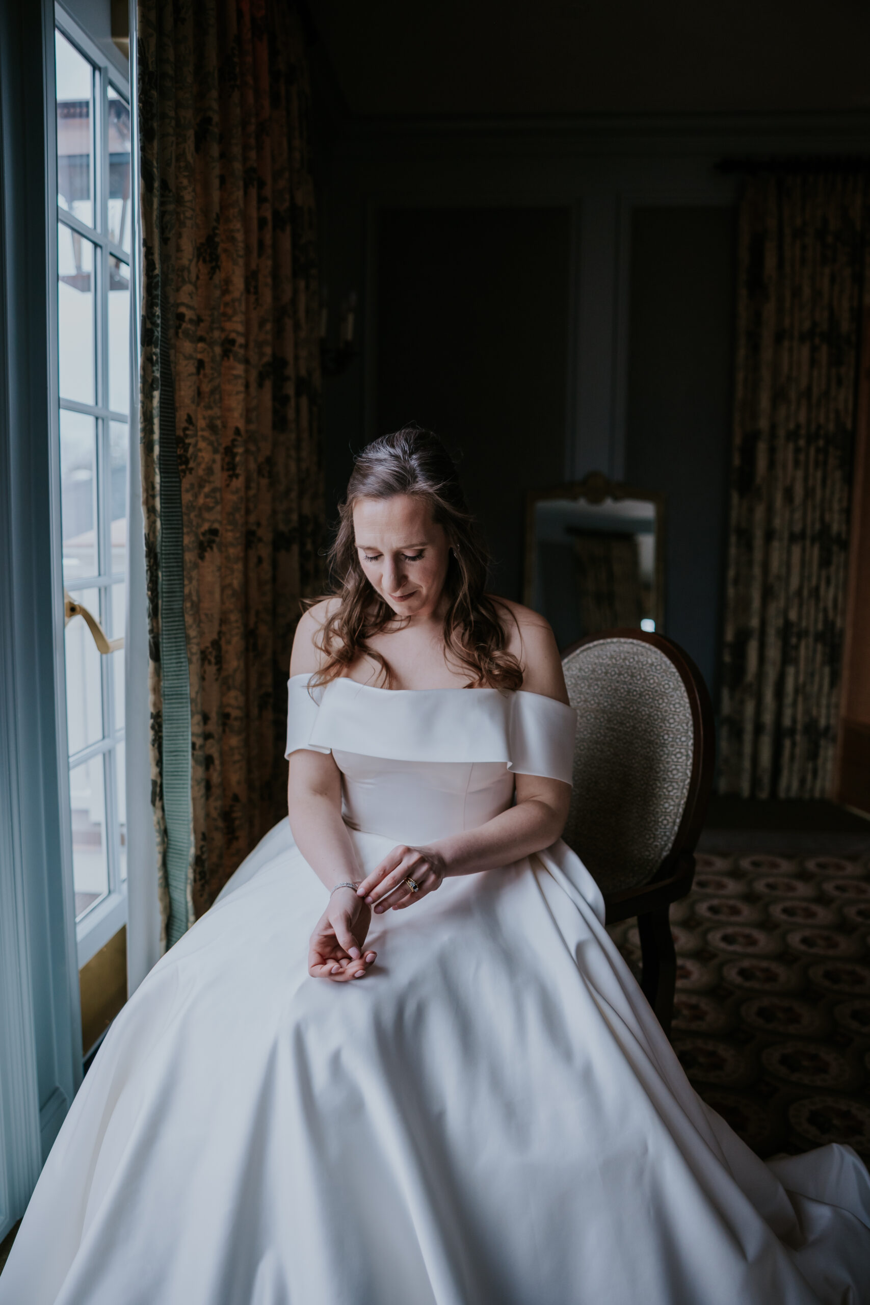 Bride in an off-the-shoulder gown quietly adjusts her bracelet while seated near a large window with moody natural light.