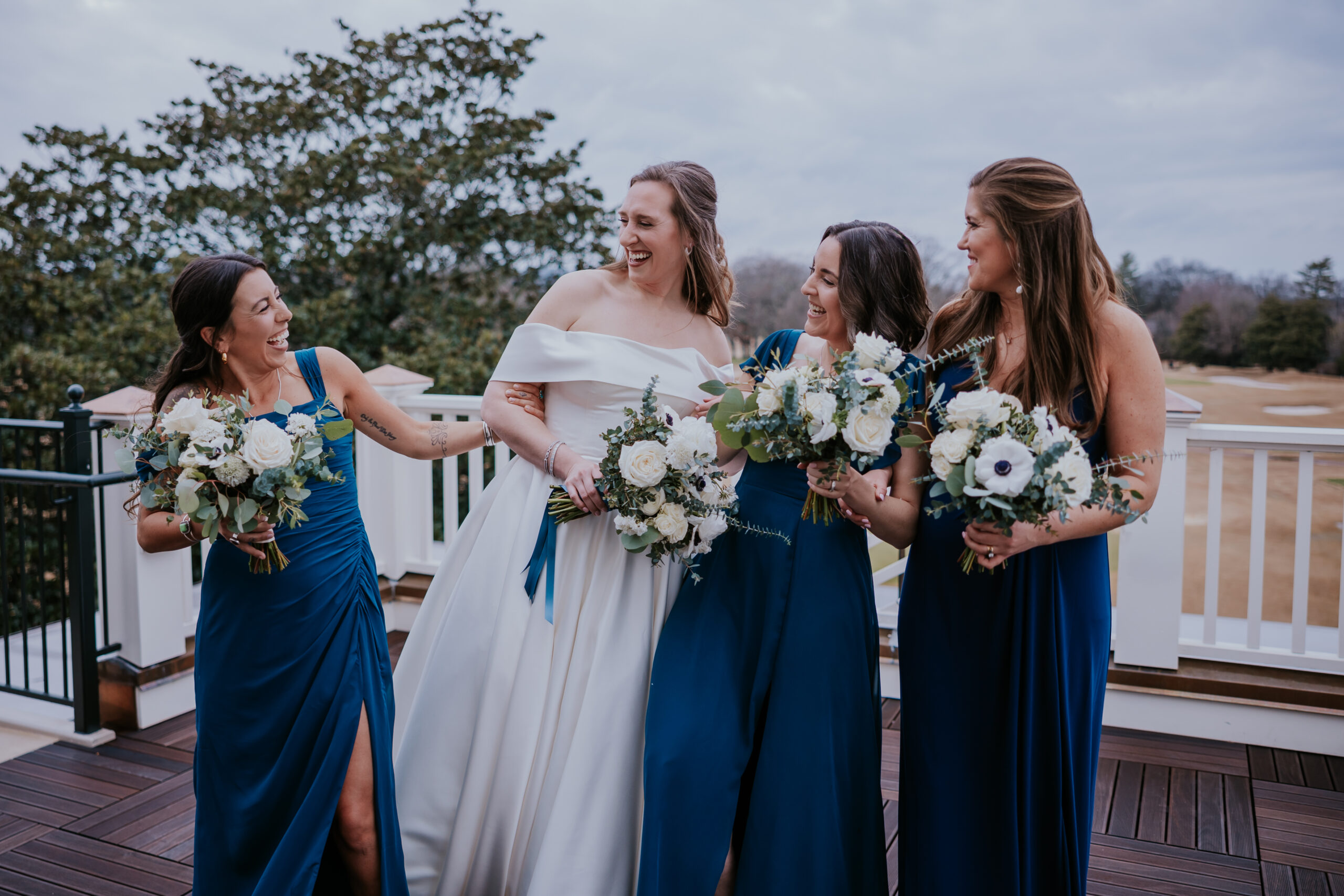 Bride and her bridesmaids share a laugh on a scenic deck, holding bouquets of white flowers and navy blue dresses complementing the mood.