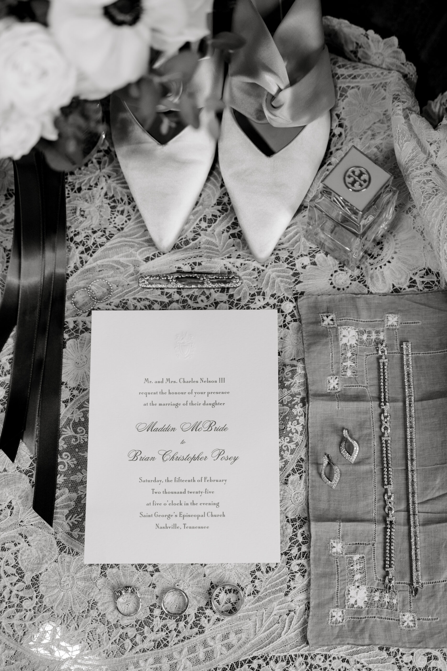 Elegant black-and-white flatlay featuring the couple's wedding invitation, heirloom jewelry, perfume, and bridal shoes on lace fabric.