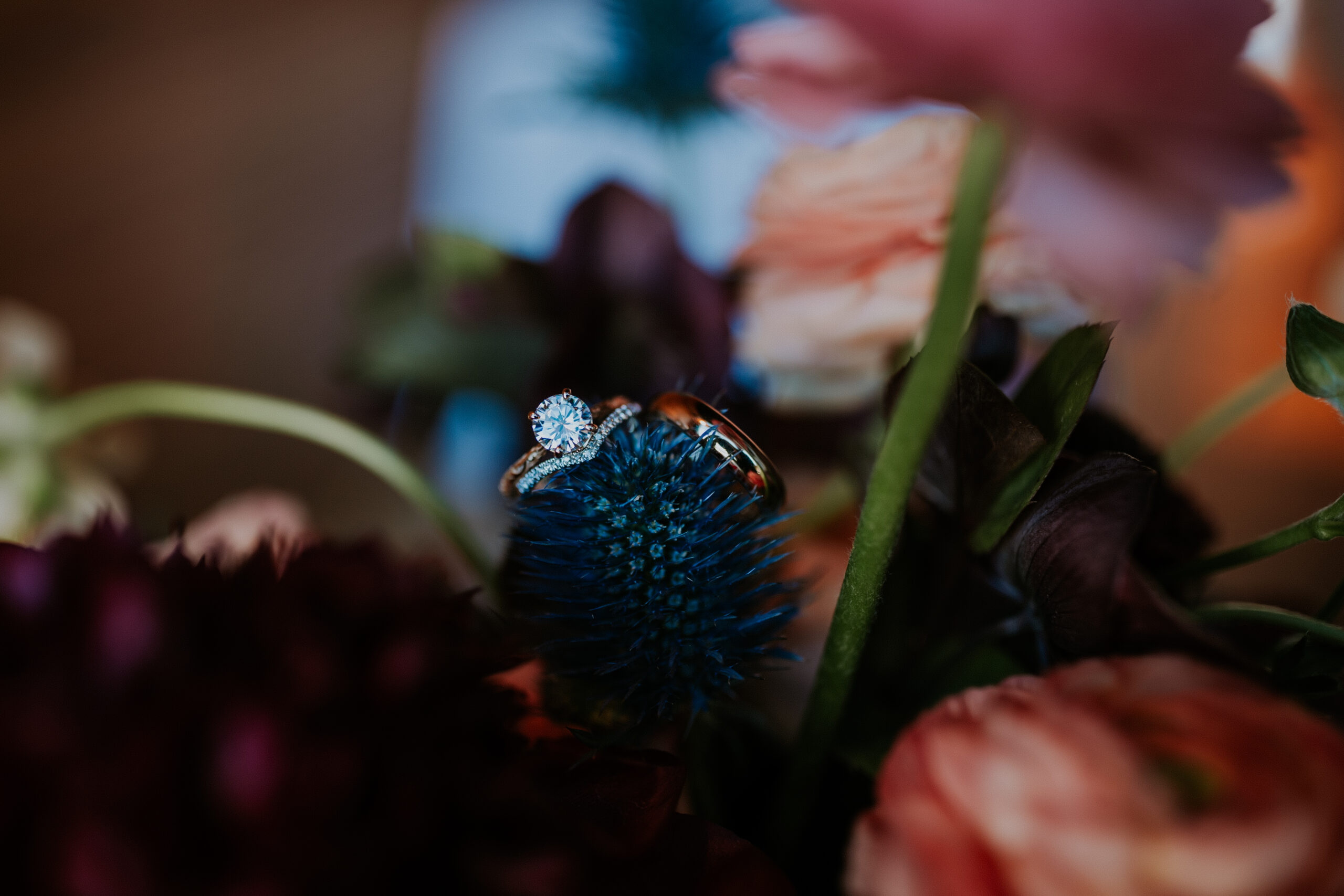 The wedding rings rest on a deep blue thistle flower, nestled among romantic blooms in the bridal bouquet.