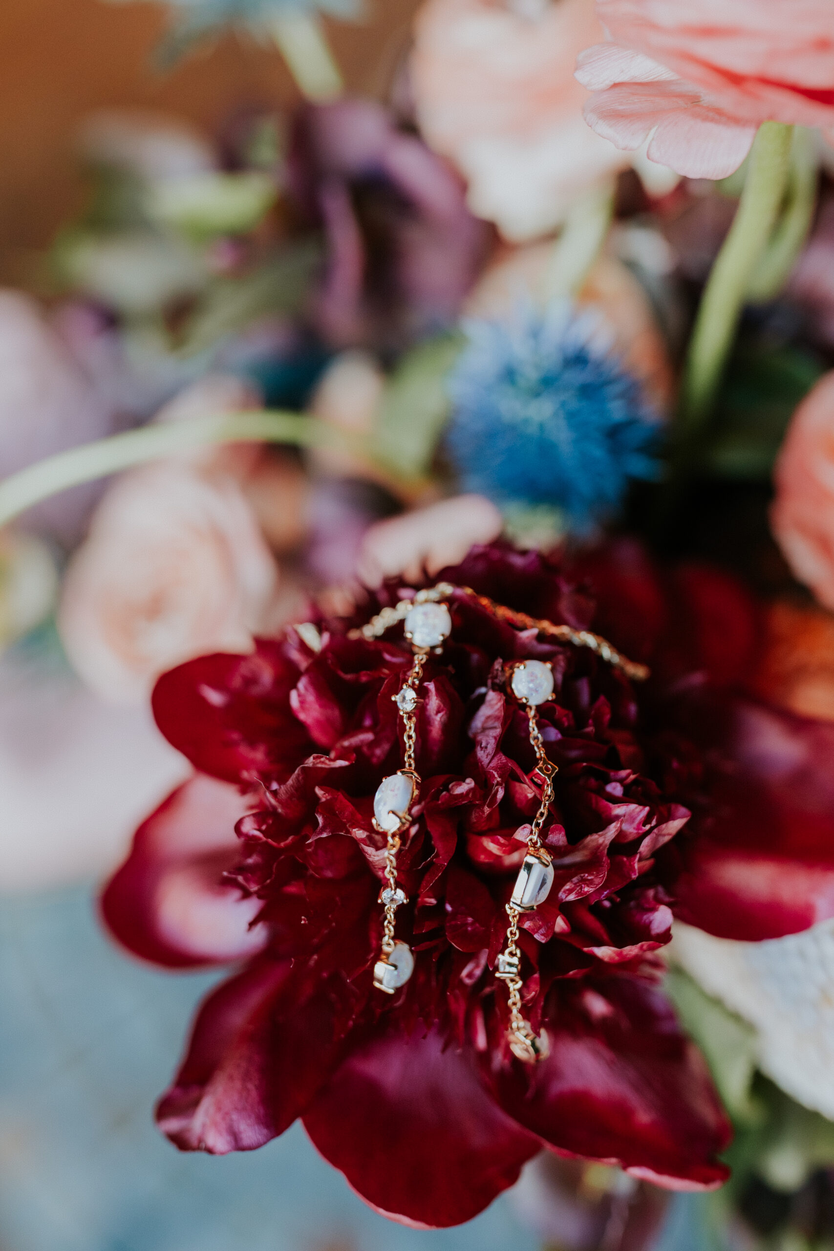 Elegant drop earrings are styled atop a burgundy peony, surrounded by soft pastel florals in the bridal bouquet.