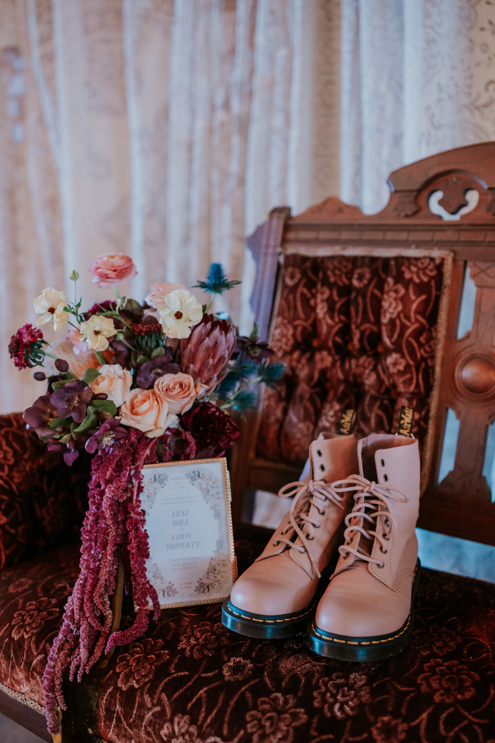 The bride’s bouquet, pink boots, and wedding invitation rest on a vintage velvet chair at a wedding in Minnesota.