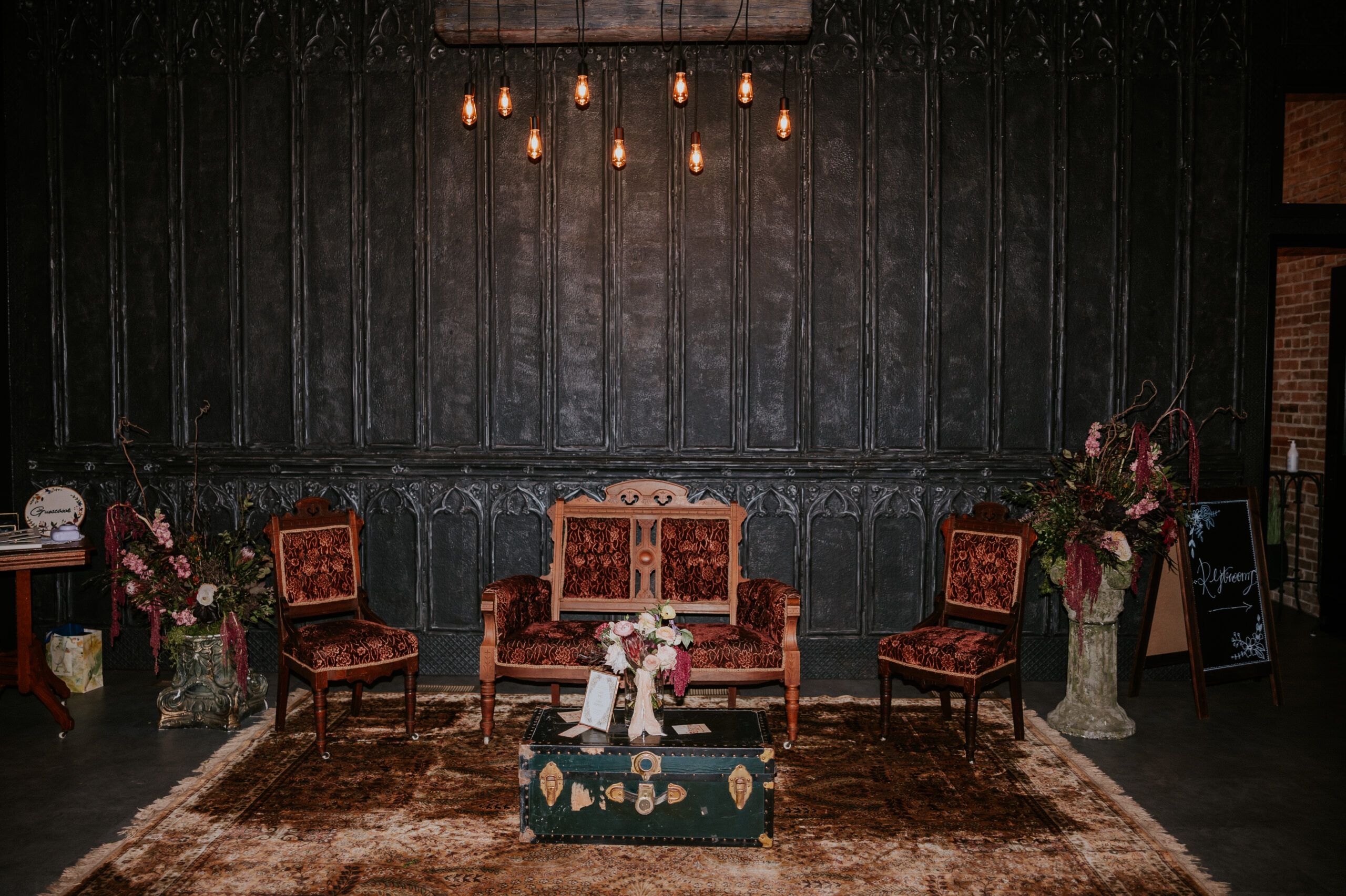 Vintage chairs and a velvet loveseat sit beneath warm Edison lights in a styled ceremony lounge area at a wedding in Minnesota.