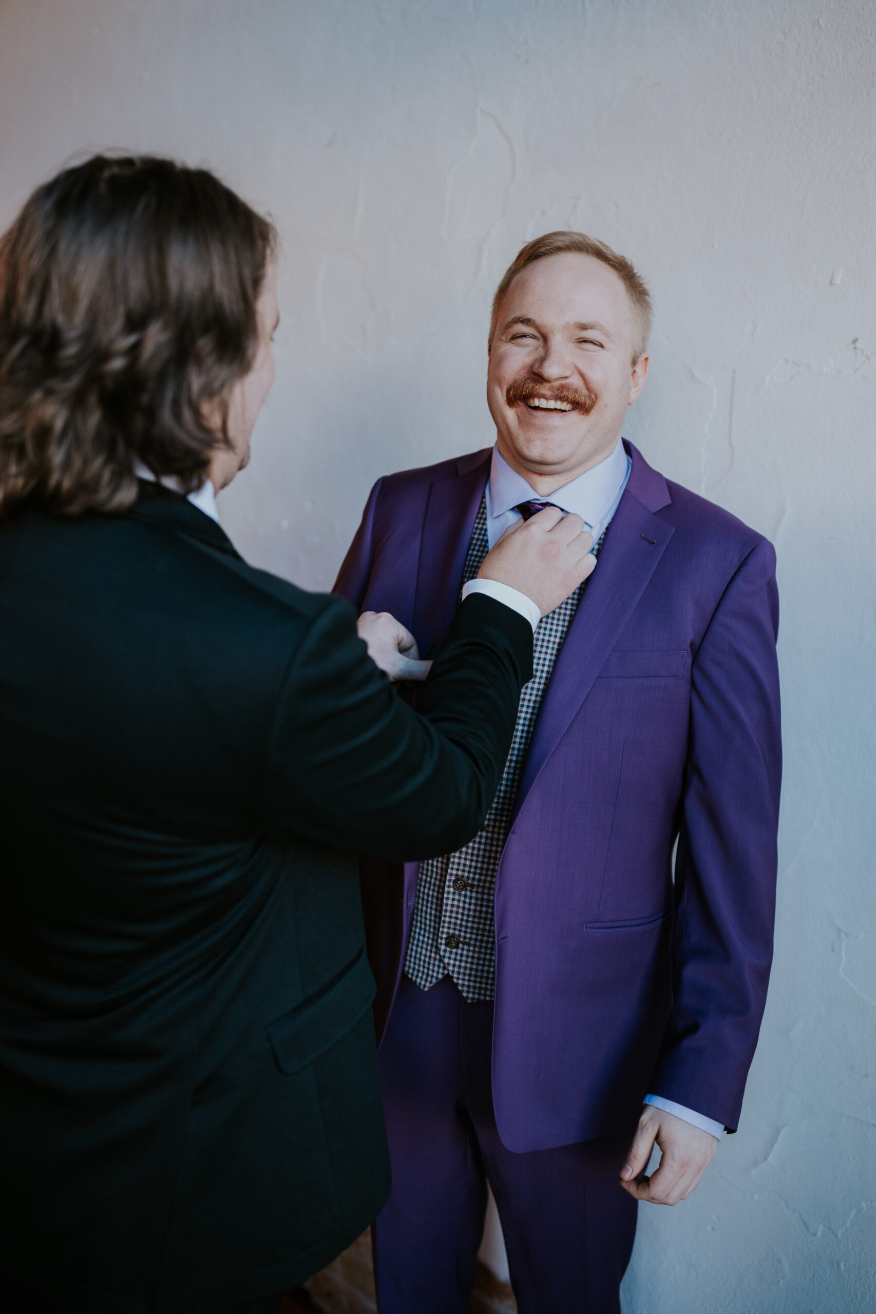 A groomsman helps the groom adjust his tie as they share a laugh together before the ceremony.