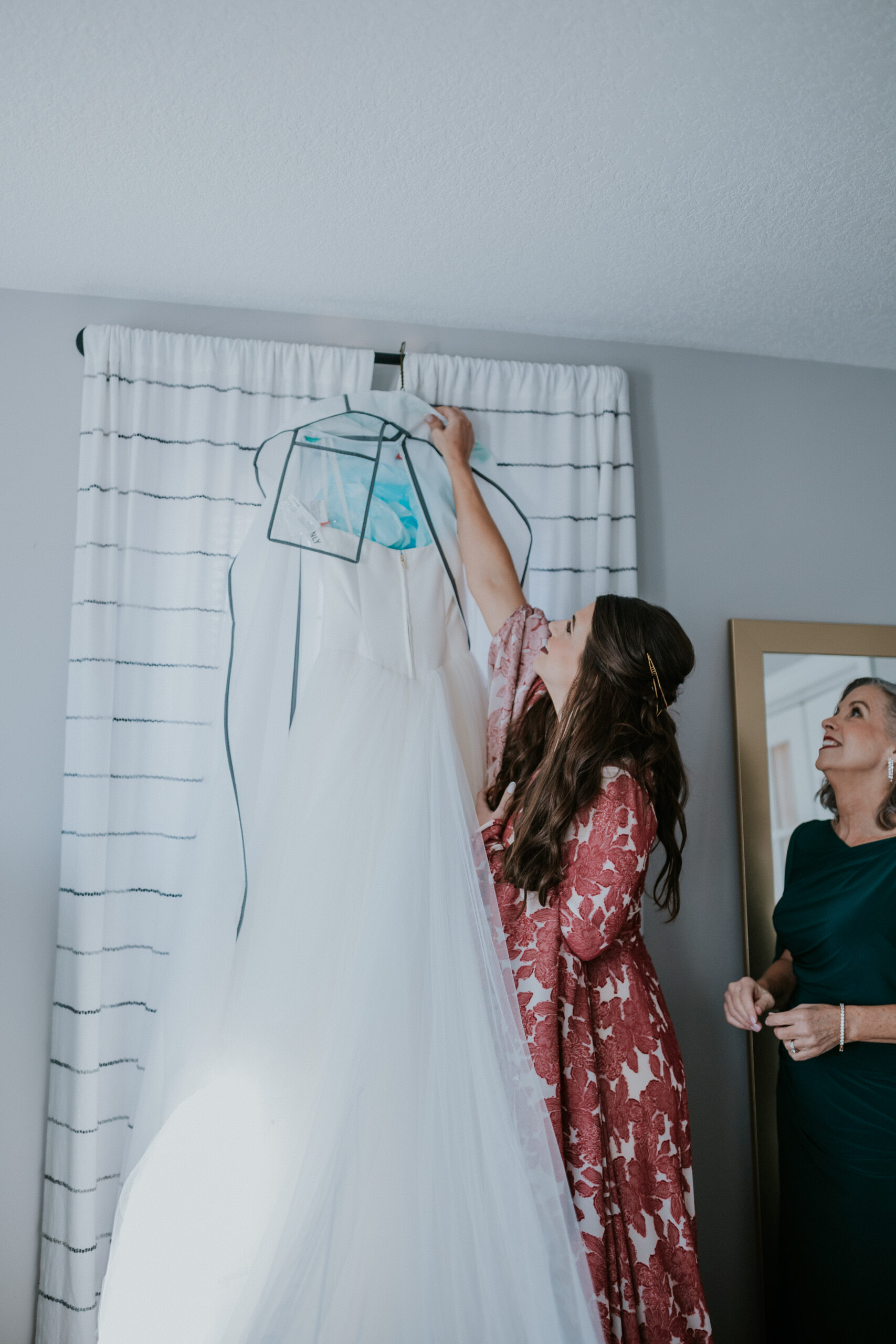 The bride and her mother reach to unhook her gown together in a soft-lit getting-ready room before the ceremony.