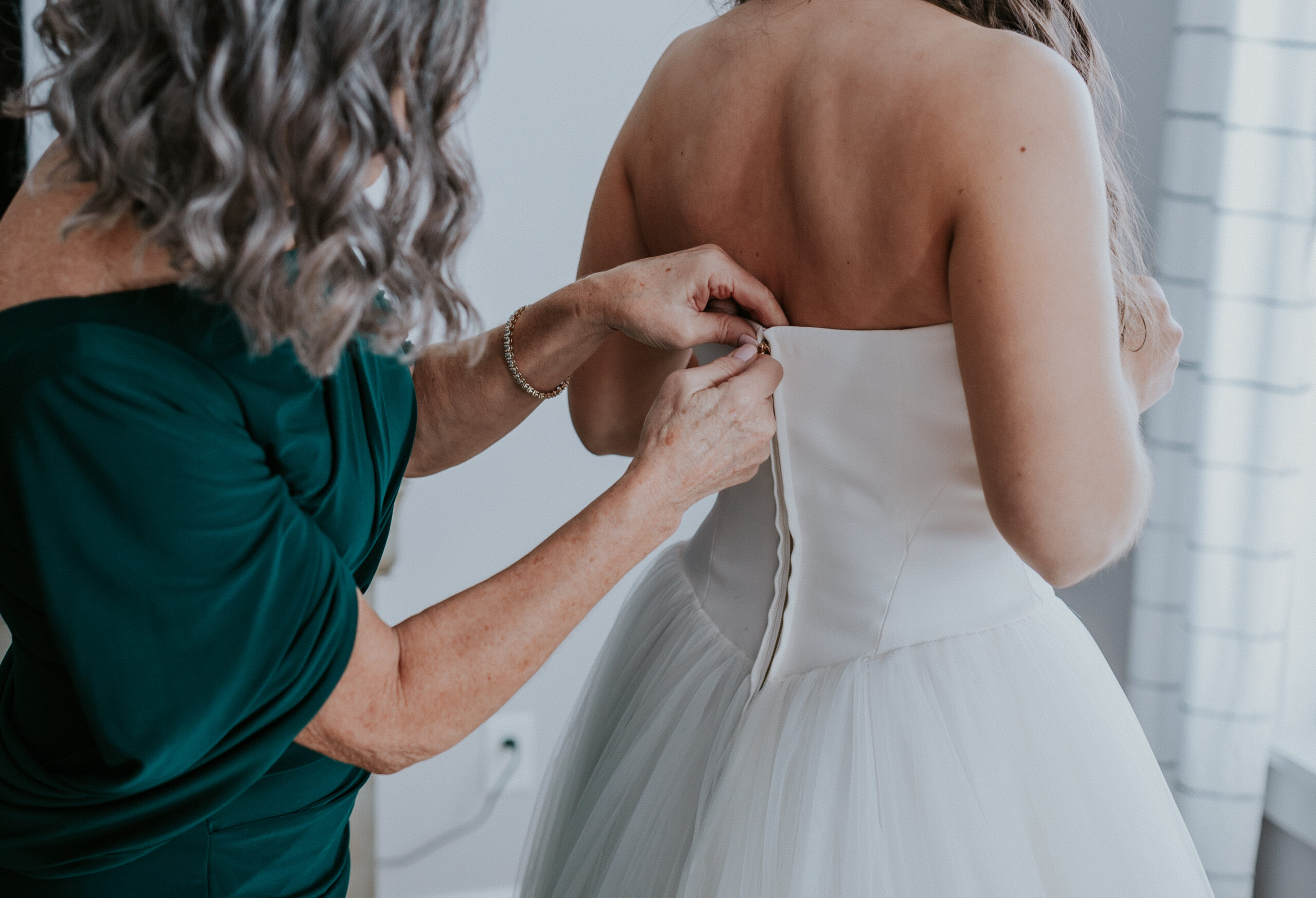 A sweet close-up of the bride’s mother fastening the back of the wedding gown, capturing a quiet pre-ceremony moment.