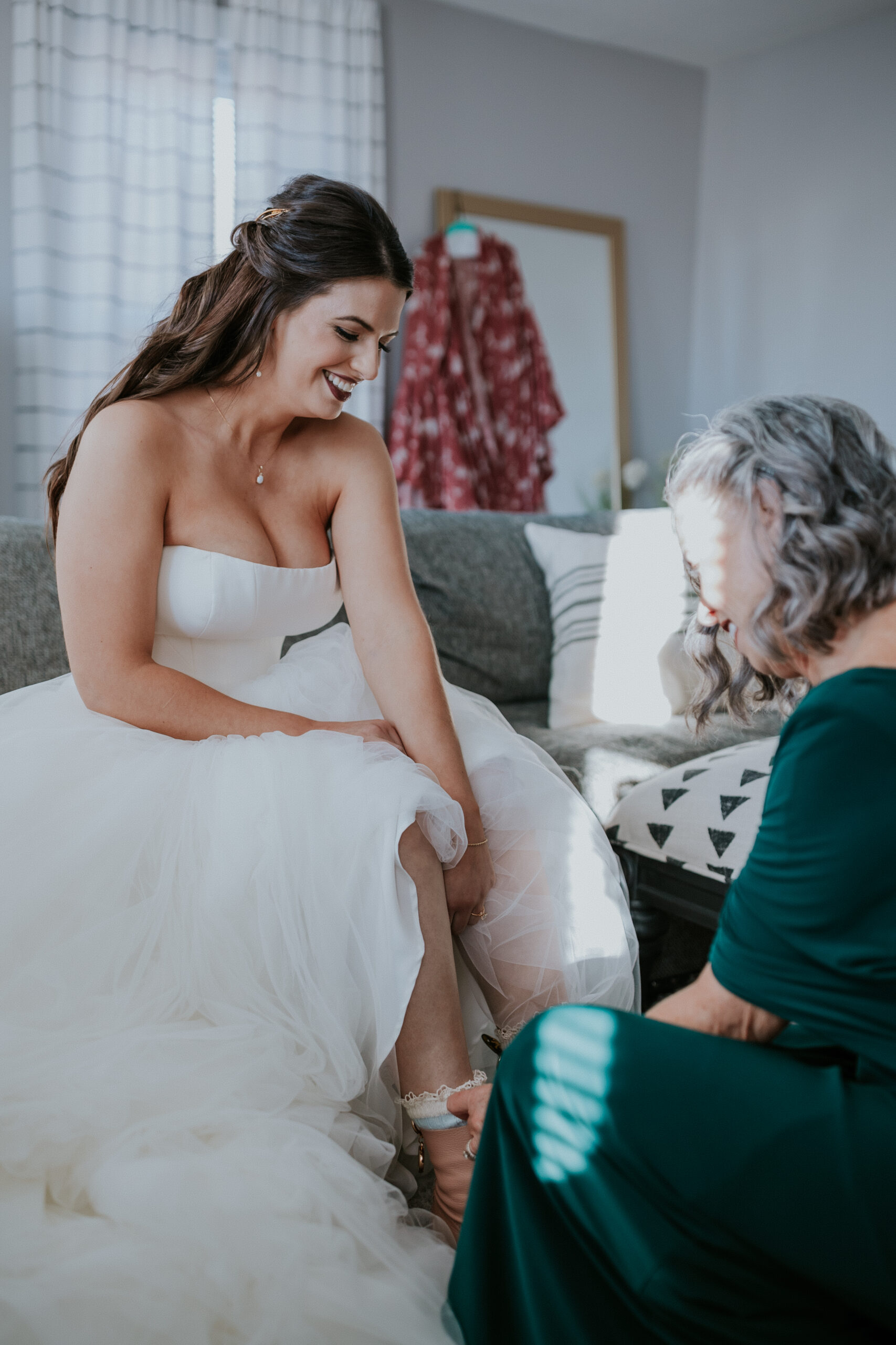 The bride smiles as her mother helps her slip on shoes, a tender getting-ready moment before the wedding begins.