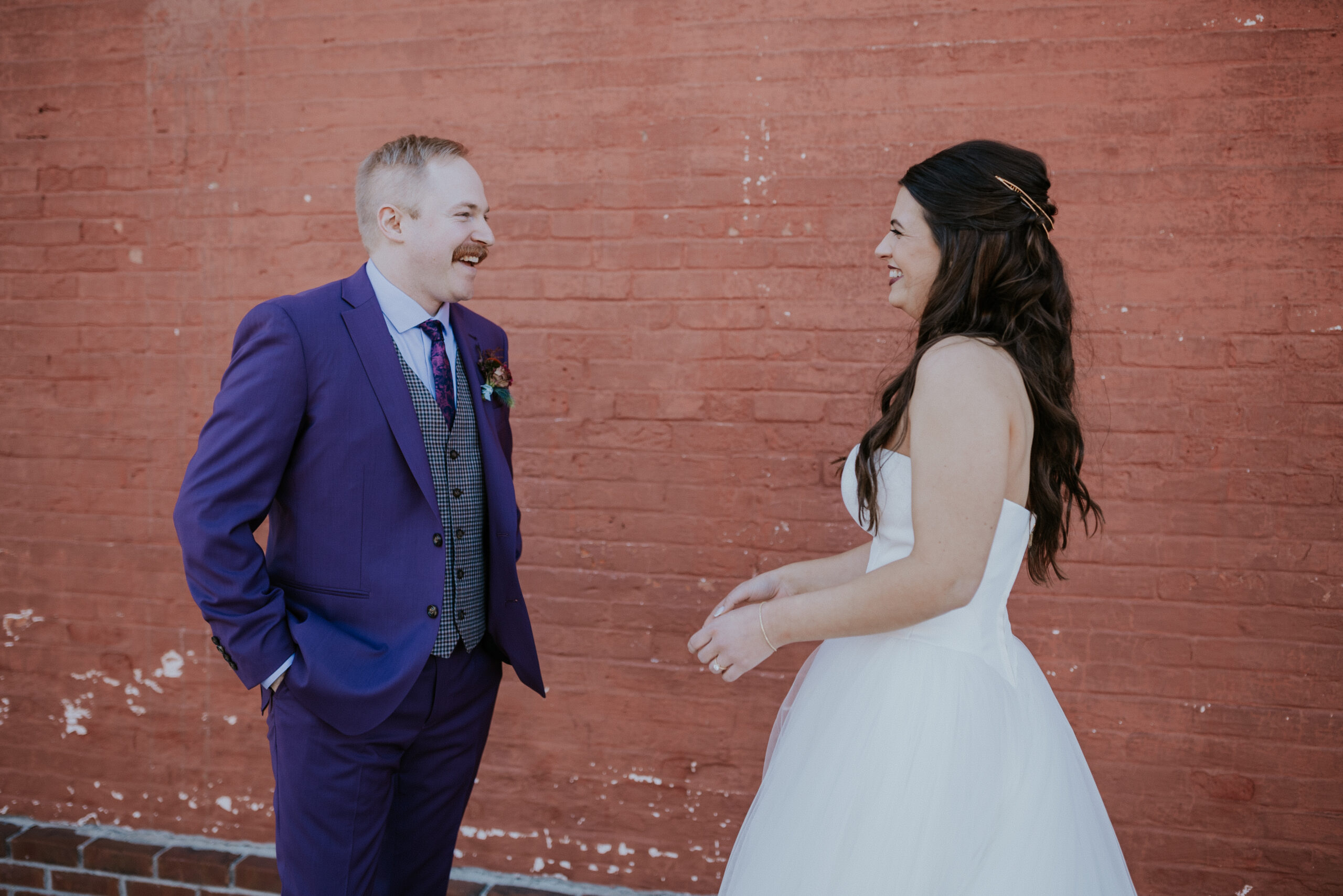 The groom beams with joy as he sees his bride for the first time in her dress, sharing a happy moment by a brick wall.