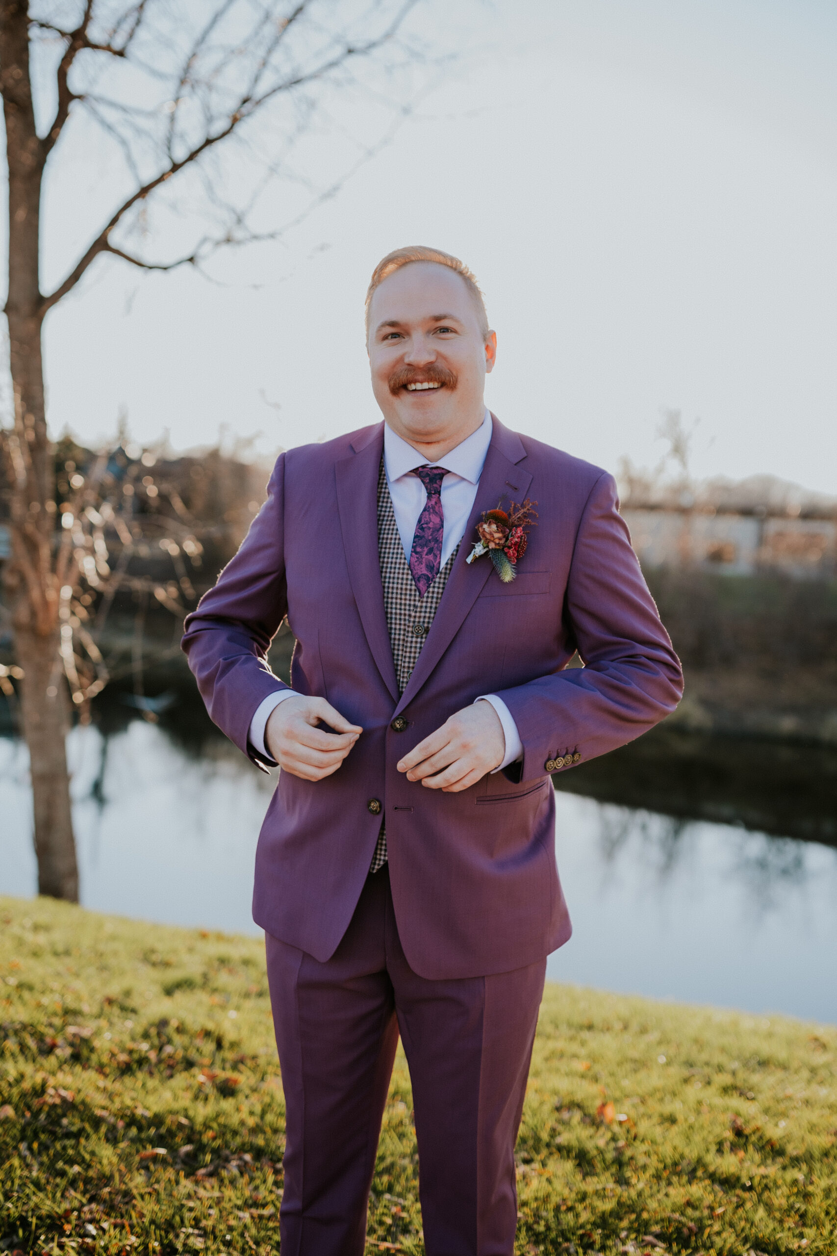 The groom smiles while buttoning his purple suit jacket, standing near the water just before his wedding in Minnesota.