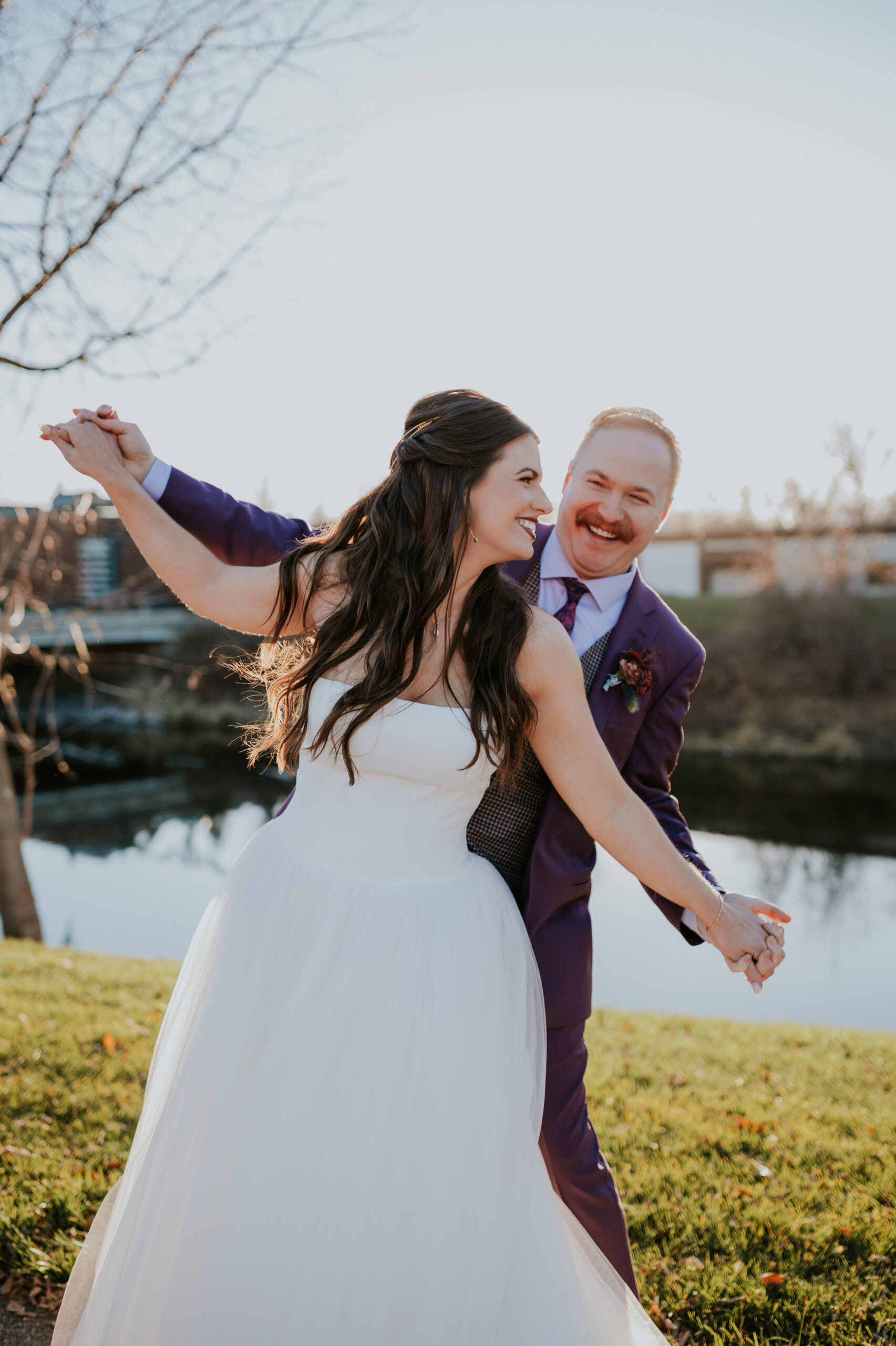 The groom dips the bride with a playful spin near the water, both laughing and full of energy on their wedding day.