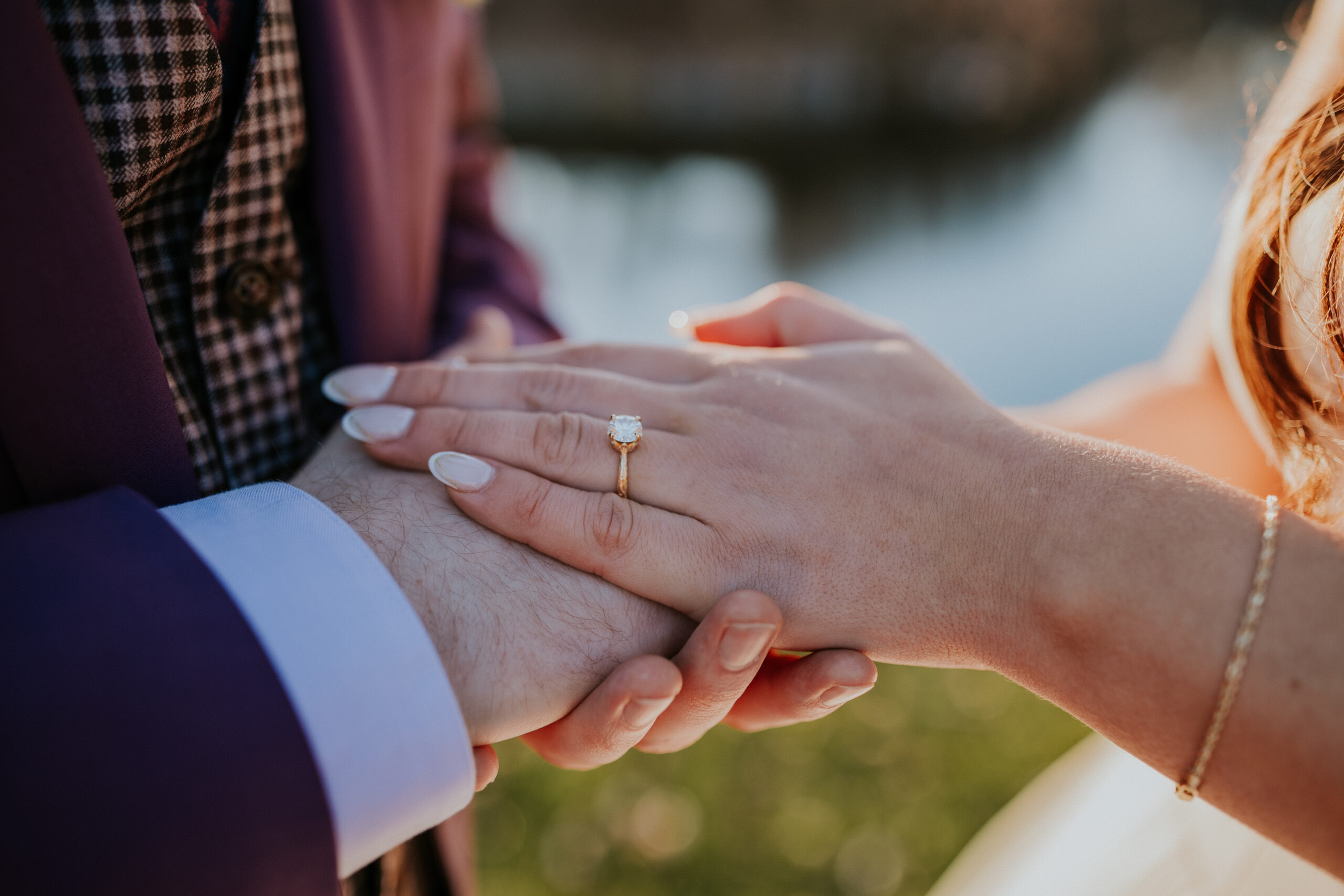 A close-up of the bride’s hand resting on the groom’s, highlighting her oval-cut engagement ring with a soft river backdrop.