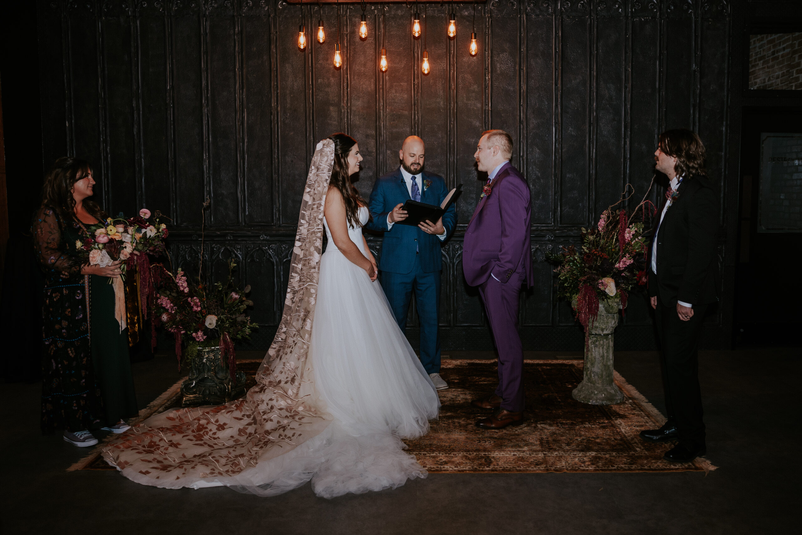 The couple stands at the altar with their officiant and wedding party, exchanging vows under warm lighting.