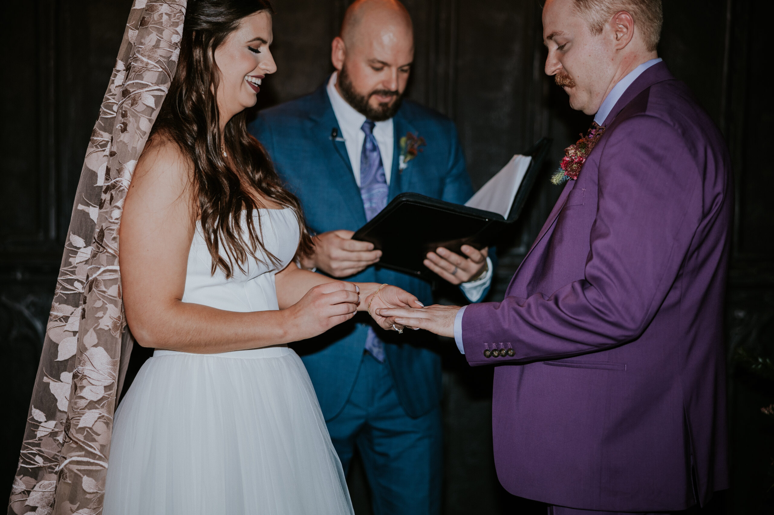 The bride places a ring on the groom’s finger during their wedding ceremony, both smiling under the officiant’s guidance.