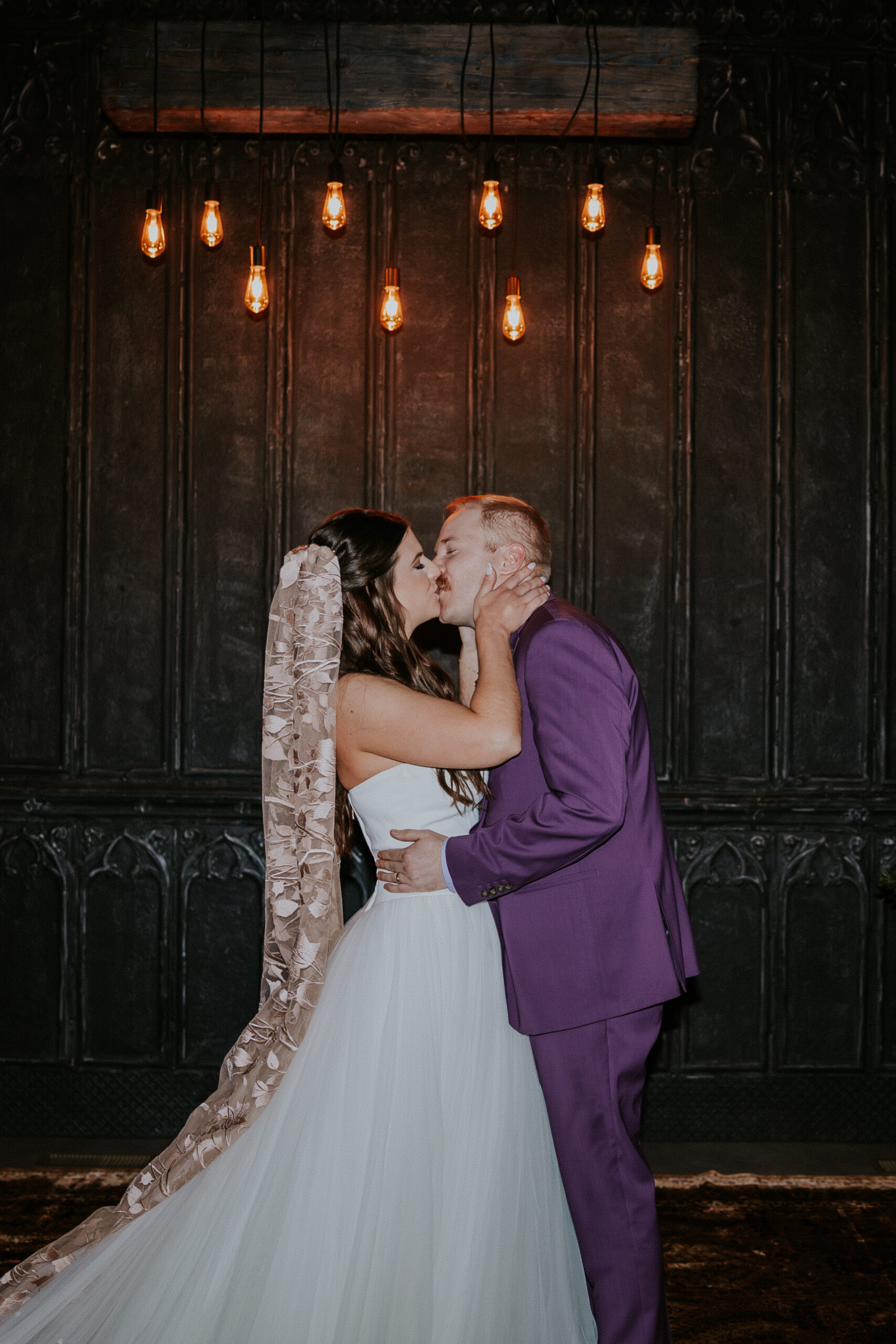 The newlyweds share their first kiss as a married couple during their wedding in Minnesota, framed by glowing Edison bulbs.