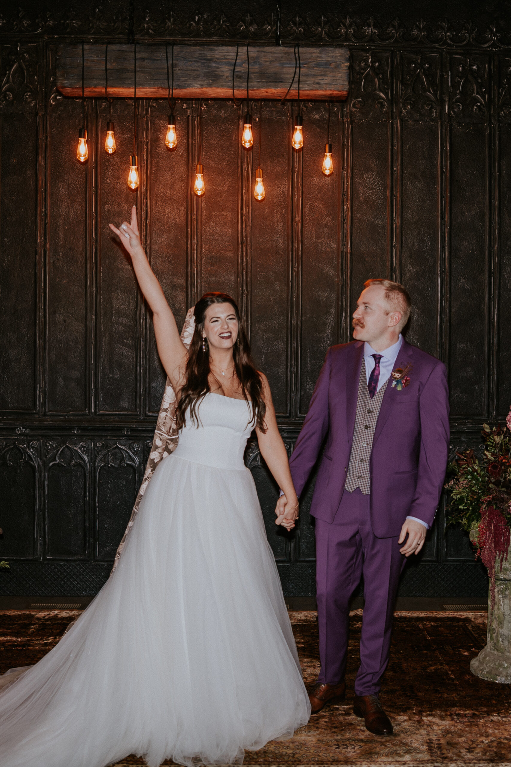The bride joyfully raises her hand in the air while holding her groom’s hand during their wedding in Minnesota, framed by warm Edison lights.