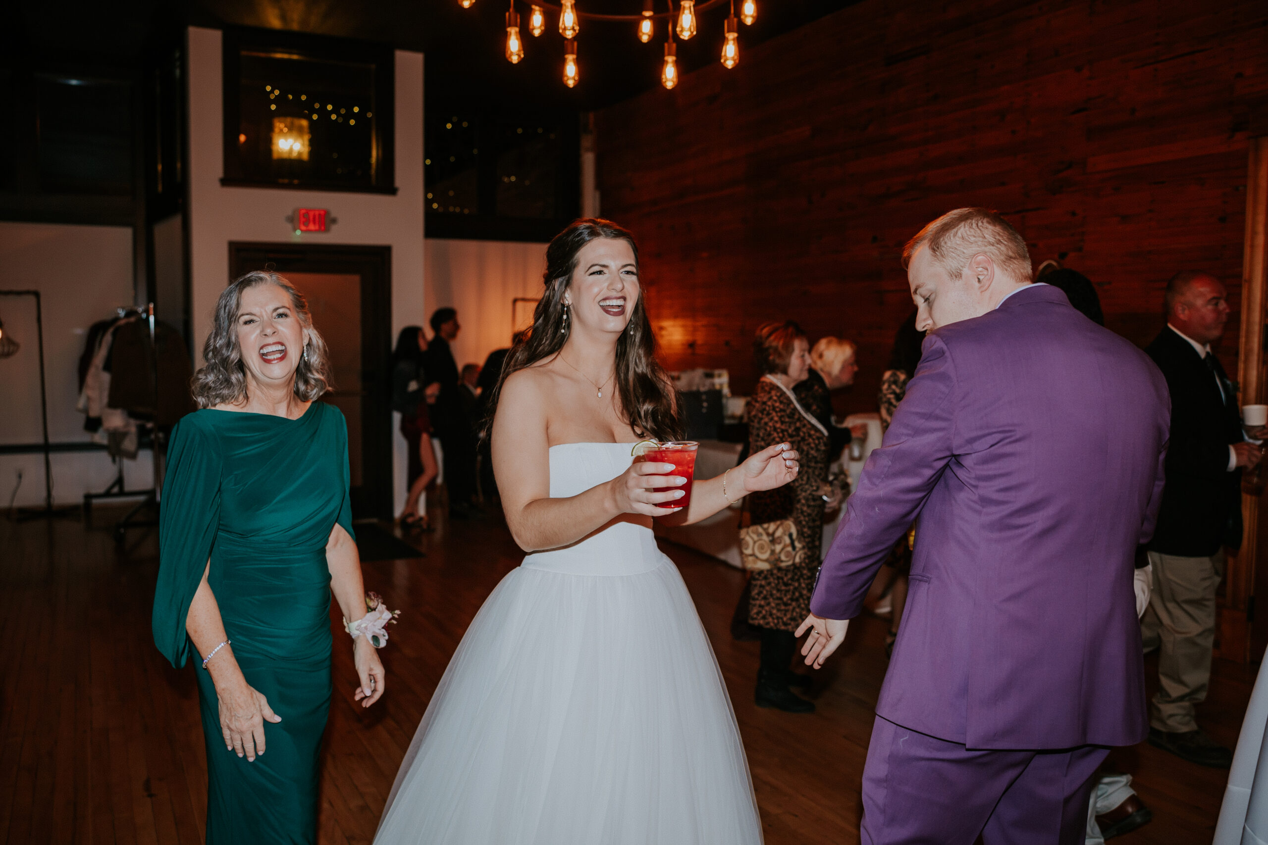The bride shares a joyful moment on the dance floor during her wedding in Minnesota, holding a drink and smiling beside the groom and a guest.